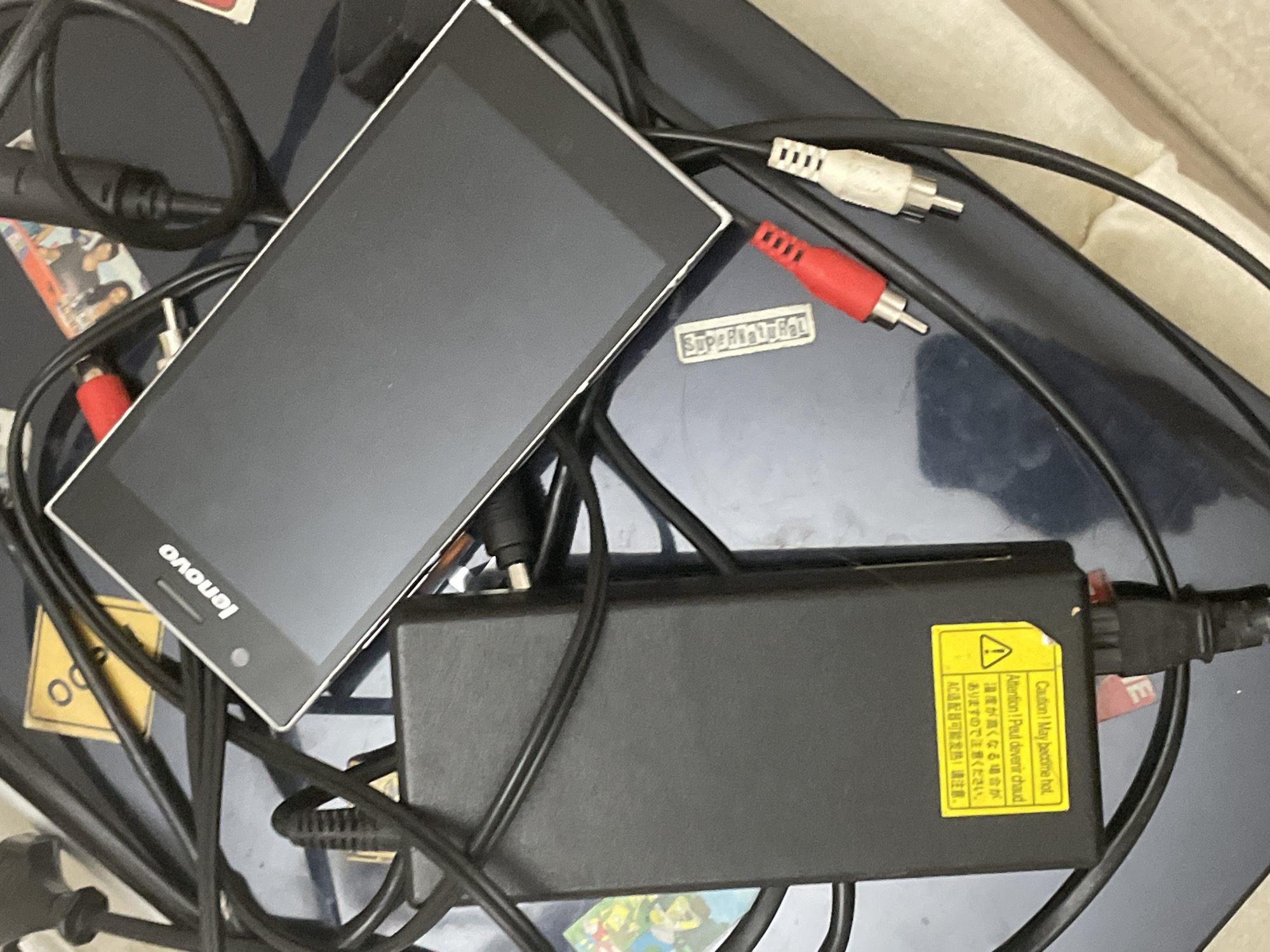The image presents a close-up, overhead view of electronic items and cables arranged on a dark blue or grey surface, possibly a desk or a large electronic device, within an indoor setting in Ihtiman, Bulgaria.
Prominently featured is a black-screened device with white edges, likely a smartphone or small tablet, displaying the "Lenovo" logo upside down. It rests diagonally across the upper left portion of the frame. Below it, a rectangular black power adapter block is visible, with several black cables entangled around and connected to it. One cable shows red and white RCA connectors.
A yellow warning label is affixed to the power adapter, featuring an exclamation mark symbol. The text on the label is multilingual, reading: "Caution! May become hot," "Attention! Peut devenir chaud," followed by Japanese characters that translate to "Temperature may become high, so please be careful," and Chinese characters stating "AC adapter may get hot! Please pay attention."
A small, silver-toned sticker bearing the word "Superficial" is visible near the center, beneath some cables. Other small, colorful items are partially obscured beneath the electronics and wires. No individuals are present in the image, and no specific activity is taking place, beyond the arrangement of these electronic components. The time of day and weather conditions are not discernible from the indoor scene.