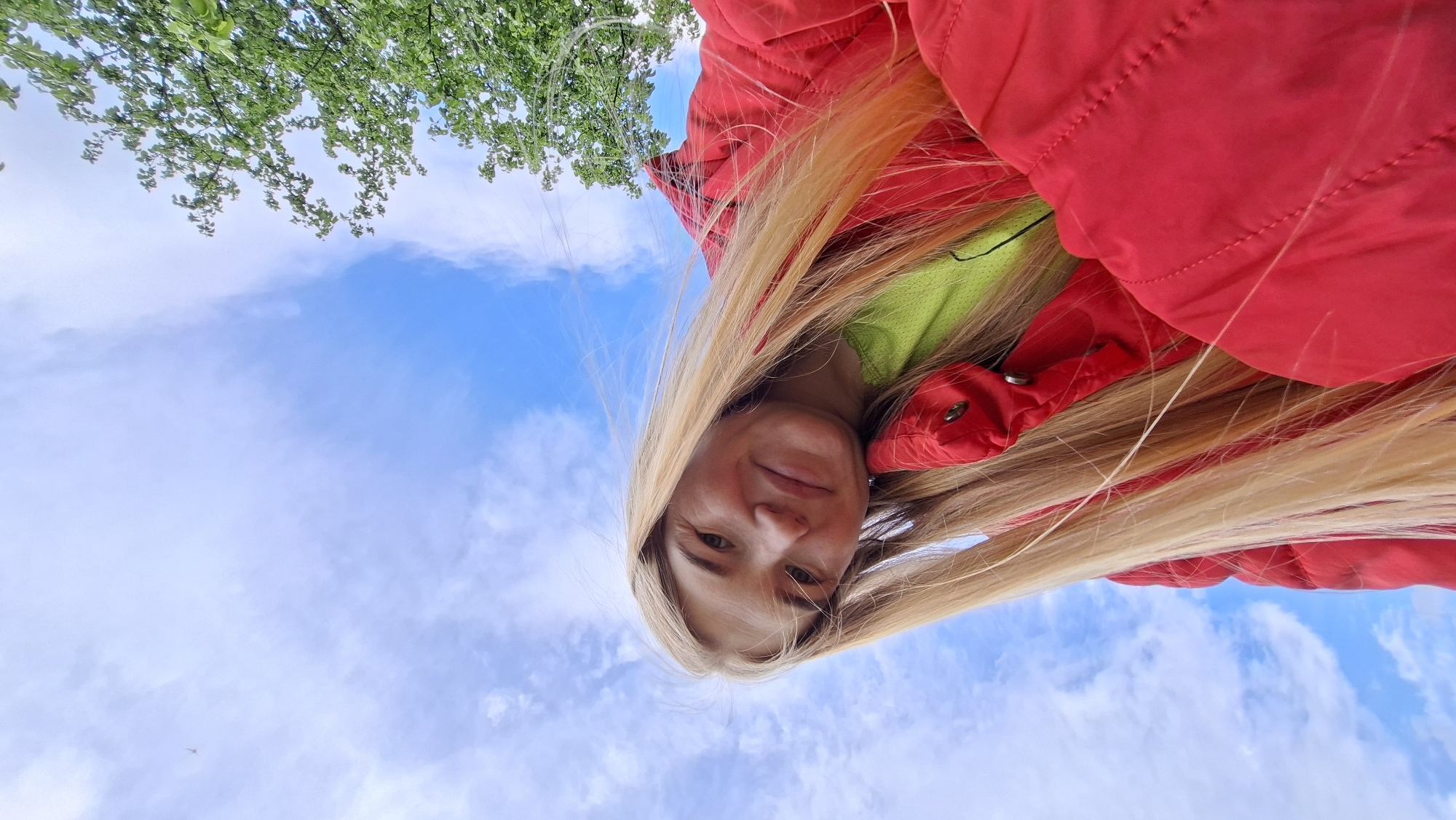 A blonde woman in a red puffer jacket and green top is pictured from a low angle against a partly cloudy blue sky with tree branches in the upper-left corner. The location is Большой Букор, Russia.