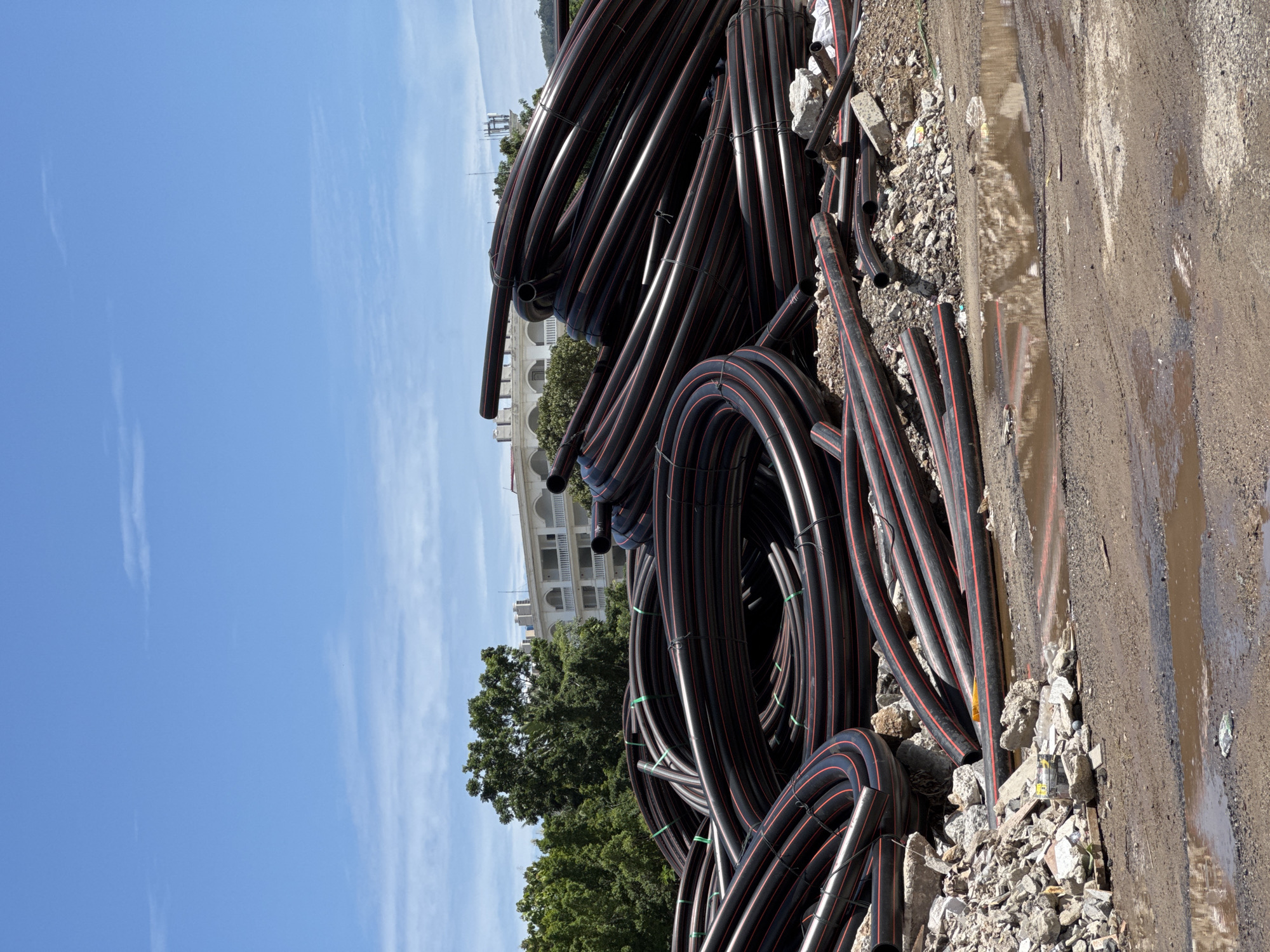 A construction site in Patong, Thailand, features a large pile of coiled black plastic pipes with red stripes. The pipes are stacked on a mound of debris, including concrete and rocks. In the background, lush green trees surround a white building with arched windows and balconies. The sky is bright blue with scattered white clouds. The foreground shows a muddy area with puddles of water, indicating recent rain or a lack of proper drainage. No people are visible in the image.