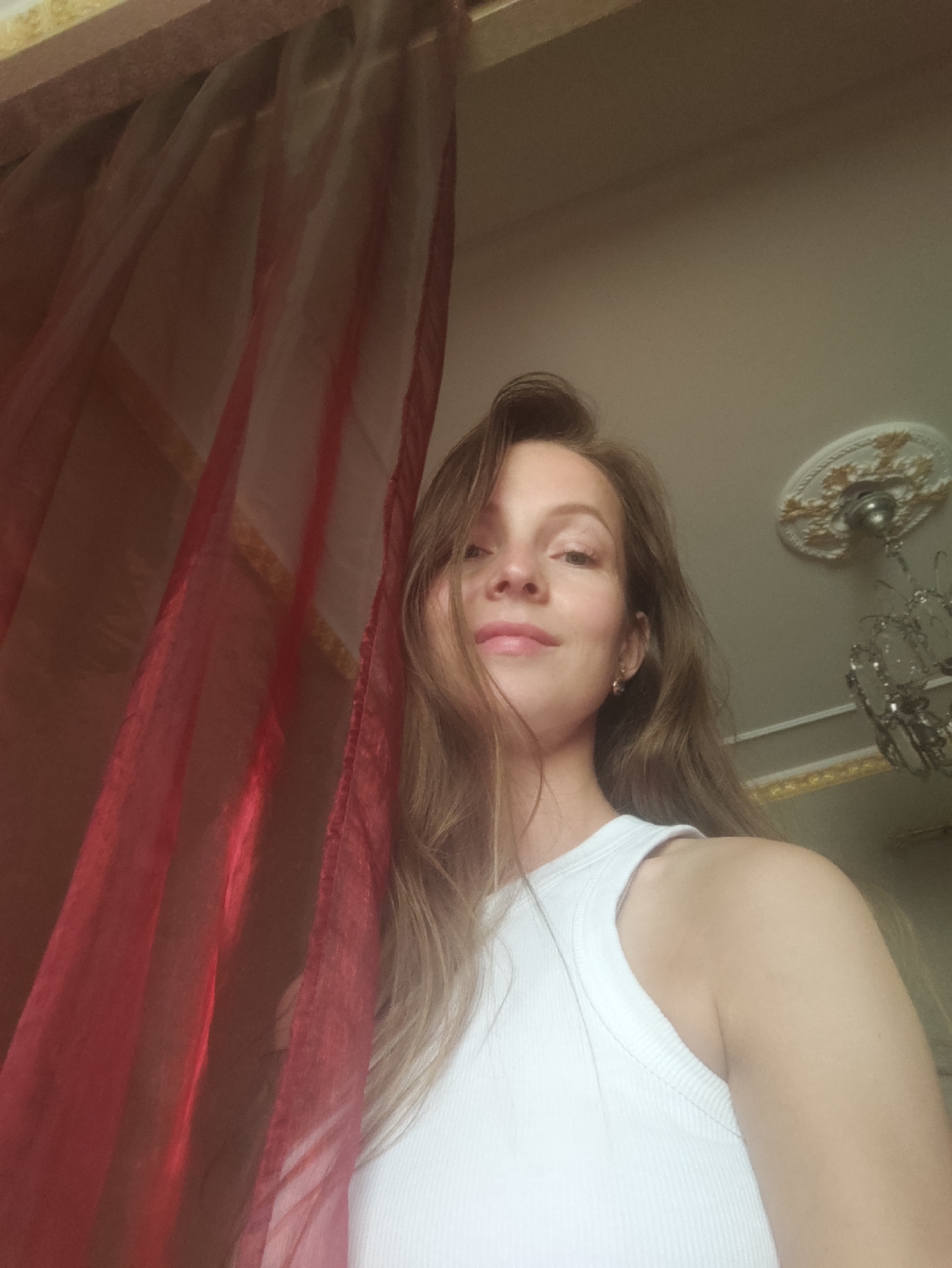 A woman with long brown hair and wearing a white tank top is in a room, possibly in Chaikovsky, Russia. A red curtain is partially obscuring the view. In the background, there is a ceiling with decorative molding and a chandelier. The photograph is taken from a low angle.