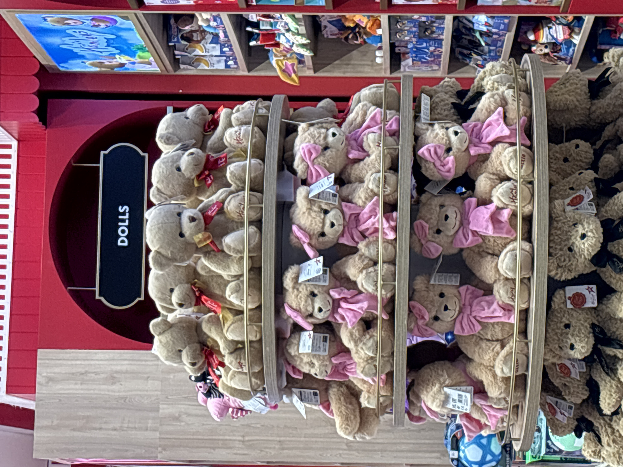 This image depicts a display of teddy bears inside a store, likely a toy store or a department store. The bears are arranged on multiple shelves, with several rows of teddy bears visible. The bears are predominantly light brown, with some wearing red or pink bows. The store features red decorative elements, including a curved backdrop with a sign that reads "DOLLS." To the right of the teddy bear display, shelves are stocked with various toys and merchandise, including what appears to be packaged games or dolls. A display screen showing animated characters is also visible in the background. The lighting suggests an indoor environment. There are no people clearly visible in the frame. The overall presentation is of a retail display aimed at children. The image does not provide any specific visual cues to confirm the location as Dubai, United Arab Emirates, but it is a plausible setting for such a retail establishment.