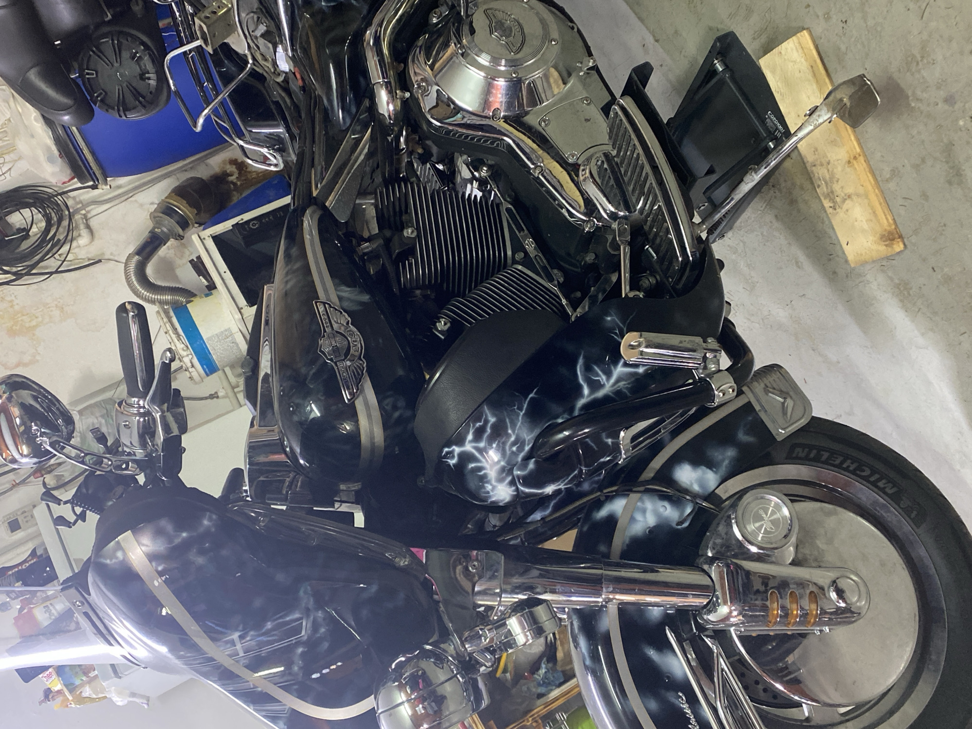 The image shows a black and chrome motorcycle with custom lightning bolt paint job parked in a garage. The garage has a light grey concrete floor. There are various items scattered around the garage, including a blue bucket, hoses, and a piece of wood. The motorcycle appears to be well-maintained and has several chrome details. The tire brand is Michelin. There is no evidence to support the location being in the city of Chaikovsky, Russia.
