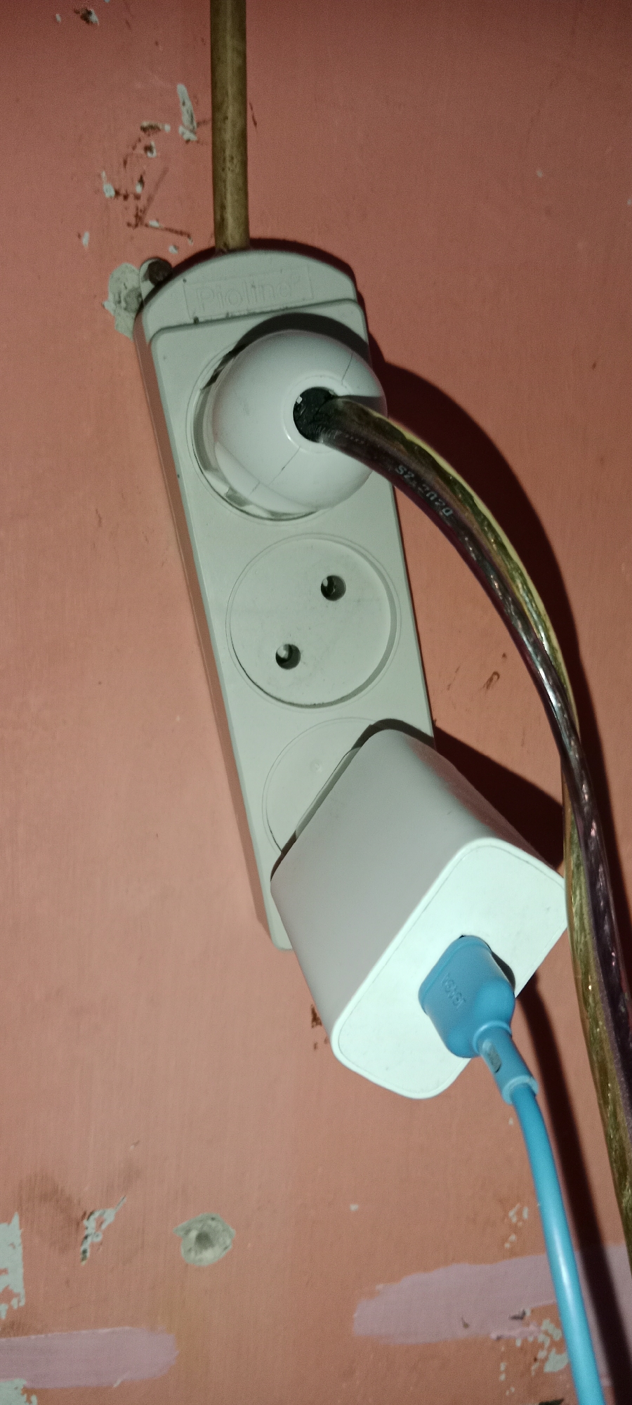 The image shows a white power strip mounted on a pink wall in Subang, Indonesia. The power strip has two outlets and a cable plugged into the top outlet. A white charger is plugged into the bottom outlet, with a blue cord extending from it. The wall appears to be peeling in some areas, with some debris and discoloration. A vertical brown pipe runs along the wall next to the power strip. The lighting suggests an indoor environment, possibly a residential or commercial space.