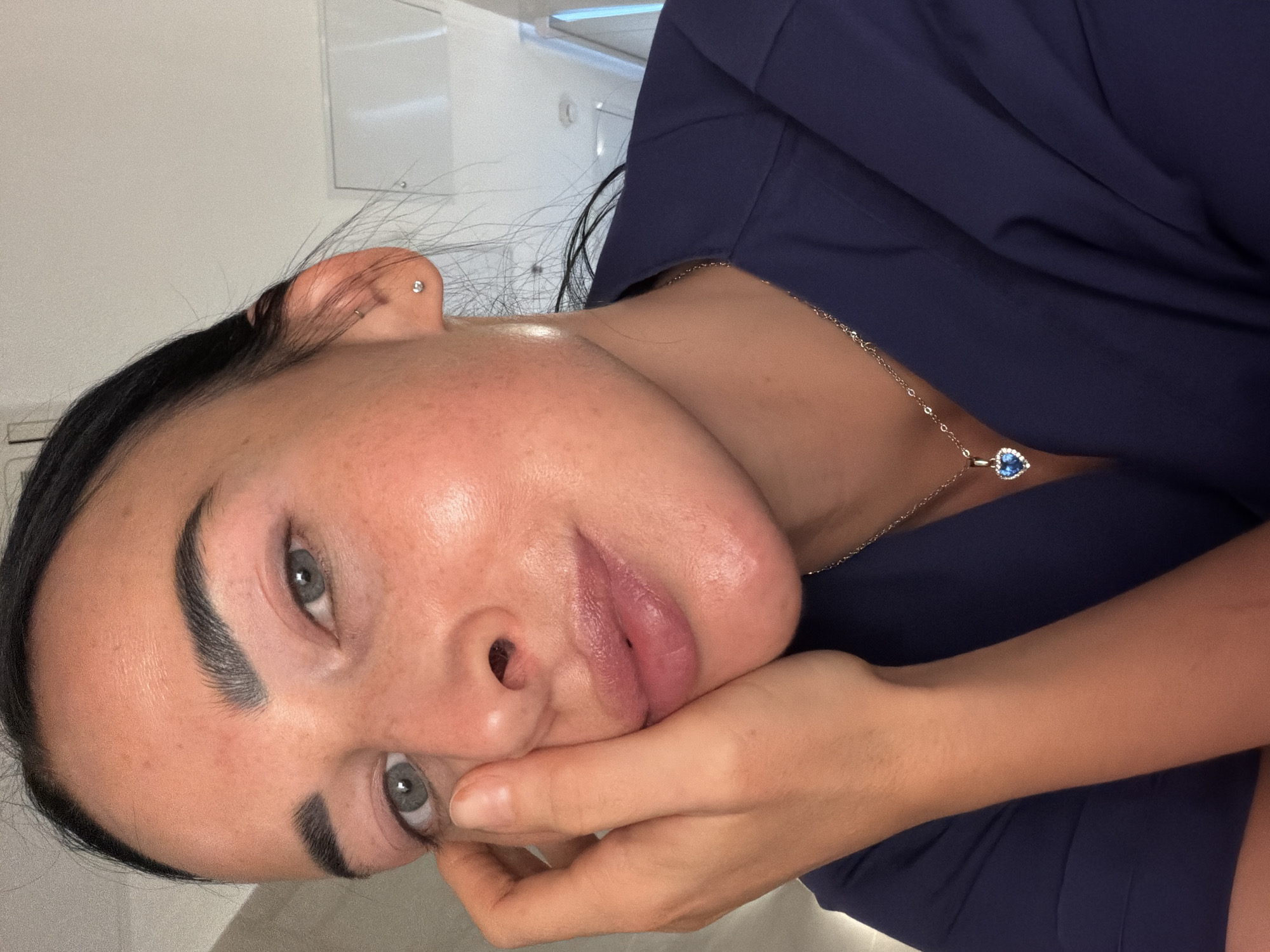 An close-up, slightly rotated portrait captures a person, likely female, with dark hair, prominent dark eyebrows, and striking light grey-blue eyes. The individual's face is central to the frame, with one hand gently resting near their chin or cheek, giving a relaxed or thoughtful pose. Their lips appear naturally full with a soft pink hue, and the skin shows natural texture.
The person is wearing a dark navy blue top or shirt. Adorning their neck is a delicate silver-toned chain necklace featuring a heart-shaped pendant with a vibrant blue gemstone encircled by smaller, sparkling clear stones. In one ear, two small silver stud earrings are visible: one in the earlobe and another helix piercing higher up on the ear cartilage.
The background is subtly lit and out of focus, consisting mainly of light-colored walls or surfaces, suggesting an indoor setting. There are faint reflections and hints of light sources, possibly artificial lighting. The overall mood is calm and contemplative. No specific location cues are visible within the image itself to confirm the provided context of Saint Petersburg, Russia, nor is there any visible text.
