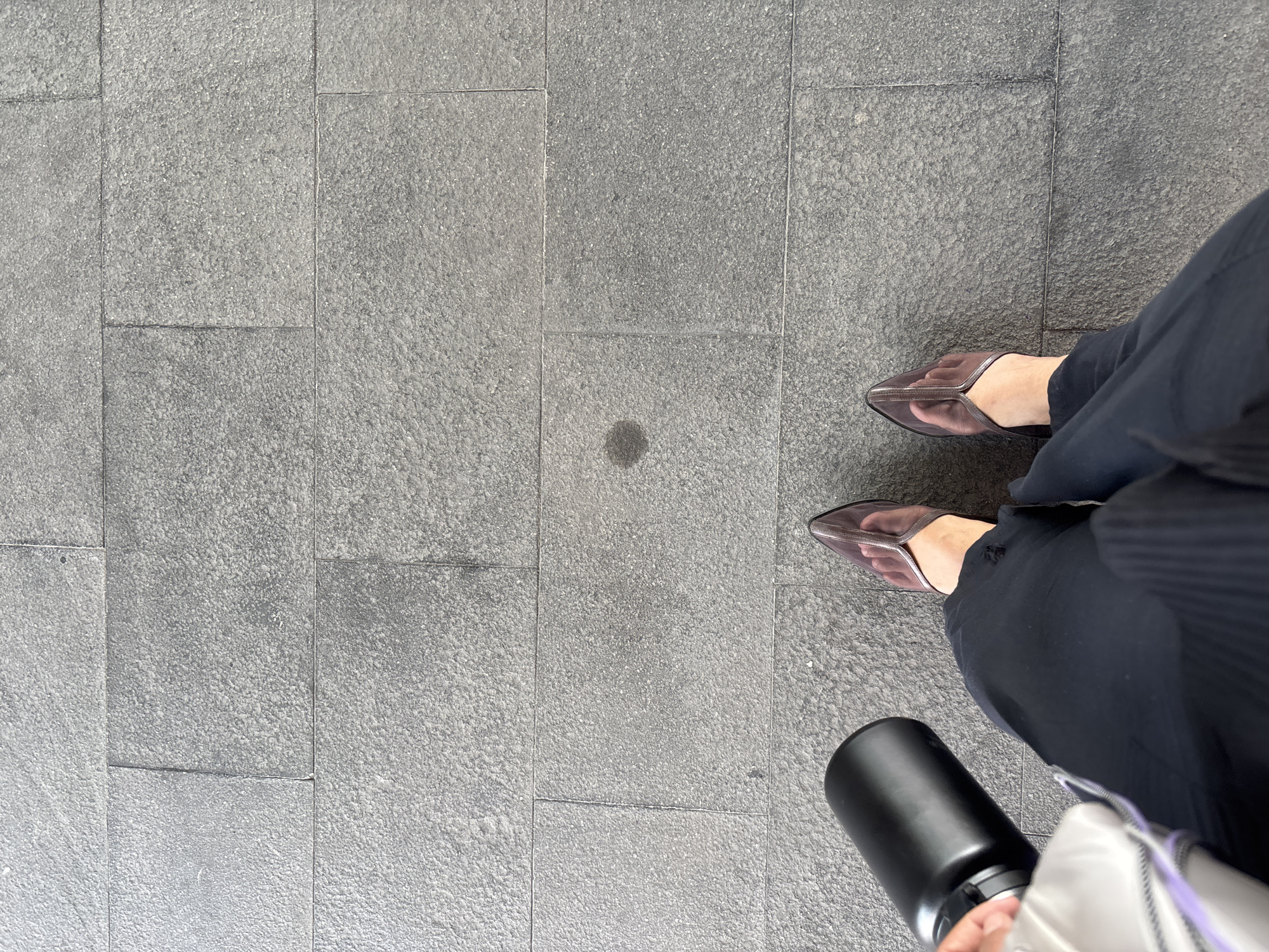A person stands on a grey tiled pavement in Binong, Indonesia. The person is wearing dark pants and sheer, pointed-toe heels. They are holding a black water bottle in their left hand and a white bag with purple accents in their right. A small dark stain is visible on the pavement between the person's feet. The tiled surface is made up of large, rectangular grey tiles, some of which show minor imperfections.