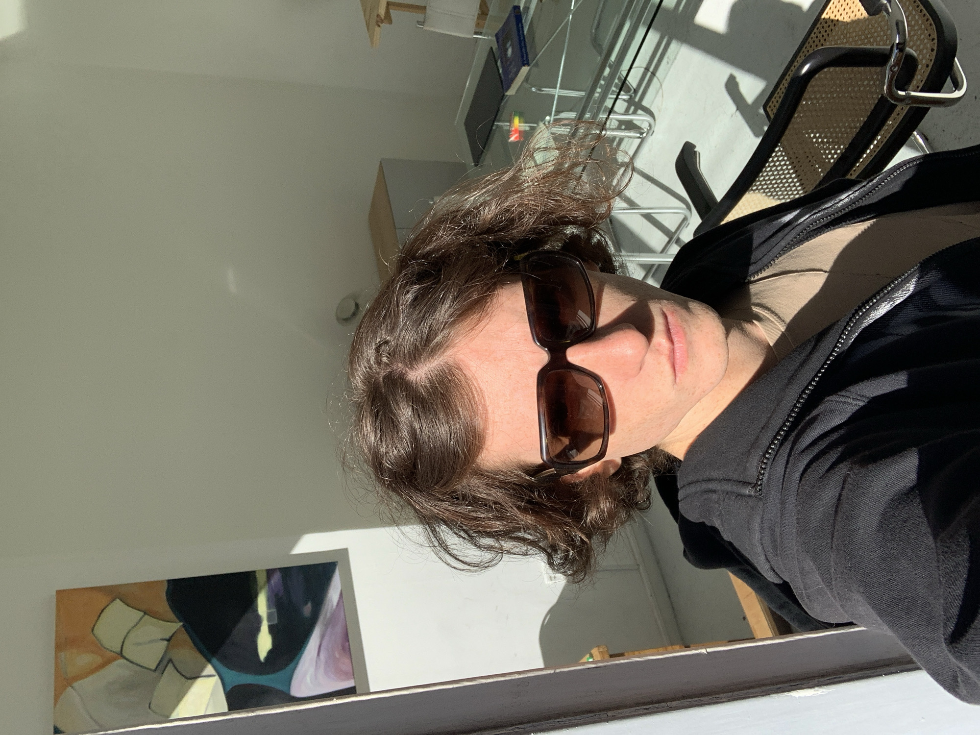 The image displays a close-up selfie of an adult with medium-length, wavy brown hair, looking upwards and slightly to their right. The person is wearing dark brown, rectangular-framed sunglasses and a black zipped hoodie over a light beige top. Strong natural sunlight illuminates the right side of their face, casting a distinct shadow and highlighting their features; their lips are visible with a neutral expression.
The setting is an indoor room with plain white walls, brightly lit by the sun entering from an unseen window. In the background, to the right of the person, a modern chair with a black frame and a woven cane-like seat and back is visible. Behind it, there's a metal shelving unit, a light-colored cabinet, and a book with a blue cover on a shelf. To the lower left, an abstract painting featuring shapes in orange, black, blue, and purple tones is displayed on a surface. A white or light-colored window sill or ledge with some texture runs along the very bottom edge of the frame.
The presence of strong, directional sunlight and distinct shadows indicates it is daytime, likely morning or late afternoon. The scene conveys a relaxed atmosphere in what appears to be a contemporary urban interior. No discernible text is visible in the image.