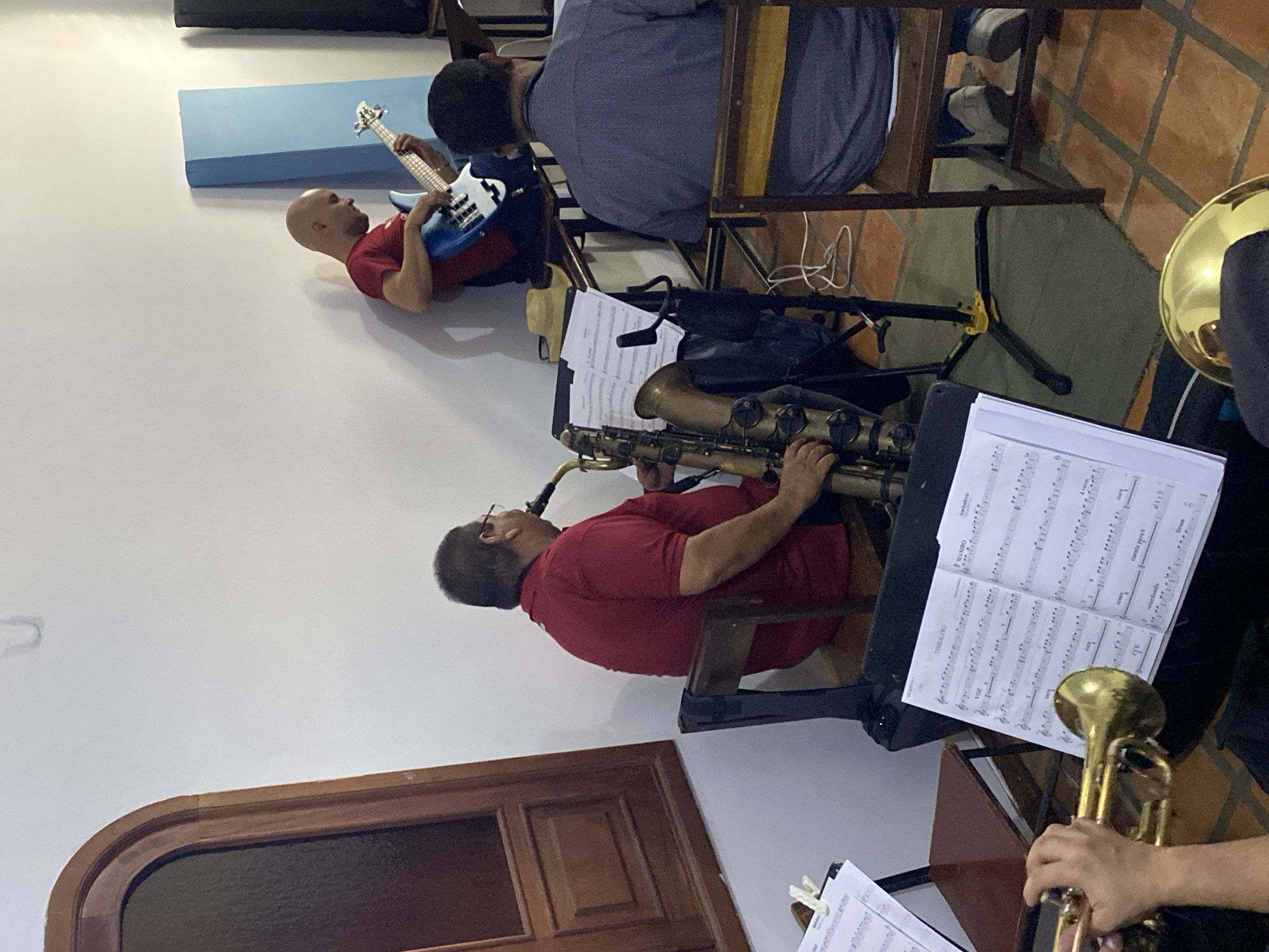 The image captures an indoor scene of several individuals engaged in playing musical instruments, likely during a rehearsal or performance. The setting appears to be a room with white walls and a reddish-brown tiled floor, possibly a community hall or practice space in Tinaquillo, Venezuela.
In the foreground, a person (whose hands are visible) holds and plays a brass trumpet, with sheet music on a stand in front of them. To the left, a man wearing a red polo shirt and glasses is seated, intently playing a tenor saxophone. Further back and to the left, another man with a bald head, also in a red shirt, is seen lying on his back on the floor while playing a blue and white bass guitar. Beside him, partially obscured by a chair, another individual with dark hair, wearing a blue patterned shirt, is seated and appears to be playing another bass guitar. A golden brass instrument, possibly a tuba or euphonium bell, is visible on the far right.
Several music stands hold sheet music, indicating a structured musical activity. Visible text on one sheet includes "TIMBALERO," "VOZ," "MAMBO," "cuenta para 6," "Drum," and "coro," suggesting a musical piece with diverse instrumentation and vocal parts, possibly Latin-influenced. The musicians appear focused on their instruments and the music. The overall atmosphere suggests a dedicated and collaborative musical endeavor.