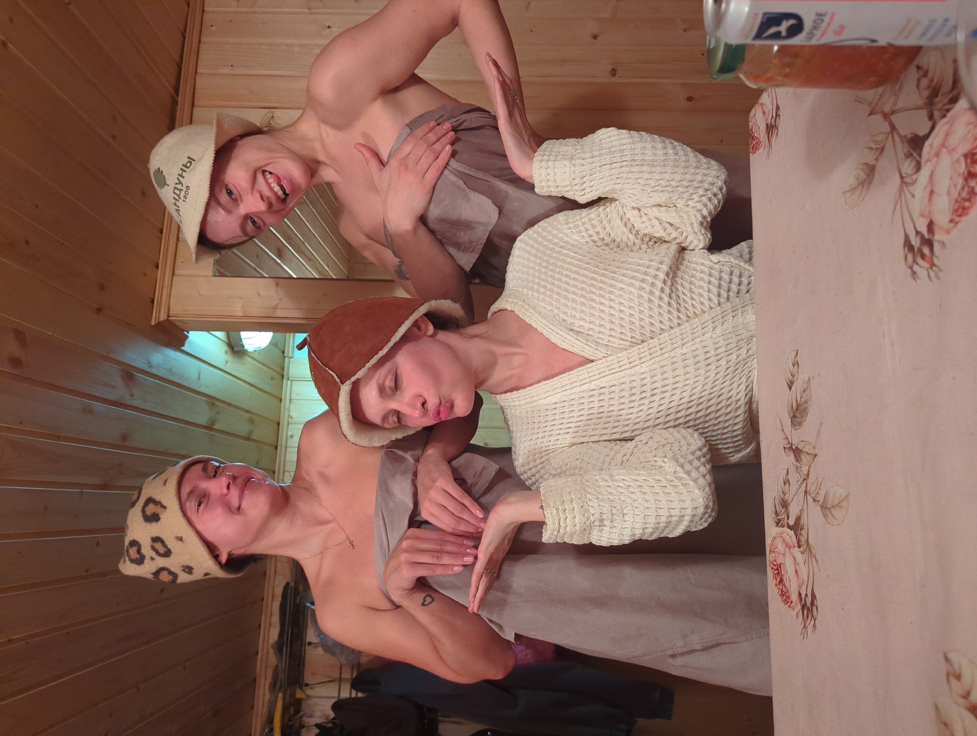 Three adult females pose in what appears to be a banya or sauna setting in Khimki, Russia. The room is characterized by light-colored wooden paneled walls and a matching ceiling.
The woman on the left wears a leopard-print felt banya hat, a light grey towel wrapped around her torso, and a gold necklace. A small heart tattoo is visible on her left forearm. She smiles towards the camera.
The woman on the right wears a light-colored felt banya hat displaying "САНДУНЫ 1808" text and a leaf logo. She also wears a light grey towel wrapped around her torso and smiles broadly with an open mouth.
The central woman wears a brown felt banya hat and a light-colored waffle-knit bathrobe. She exhibits a puckered lip expression and extends her hands forward with palms upward.
In the foreground, a table is covered with a light pink tablecloth featuring a rose floral pattern. On the table, a glass jar containing an orange substance and a partially visible silver beverage can are present. The background behind the subjects includes hanging garments on hooks and a wall-mounted mirror.