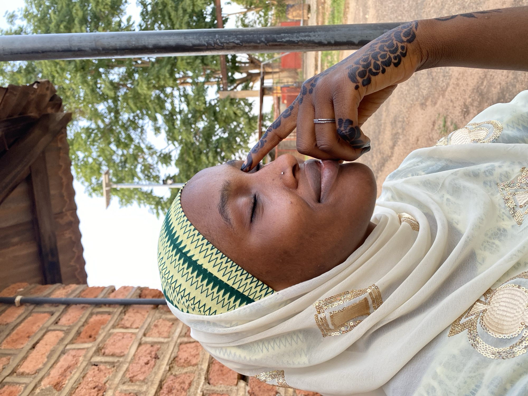A woman with dark skin, wearing a hijab with a green and yellow zigzag pattern along the forehead and a white headscarf with gold sequin embellishments, is seen in close-up. Her eyes are closed, and she has a nose piercing. Her right hand, adorned with dark henna designs and a silver ring, touches her nose. The background shows a blurred outdoor scene with a gray metal bar, trees, and possibly a red brick building. Based on the prompt, the location is Vom, Nigeria.