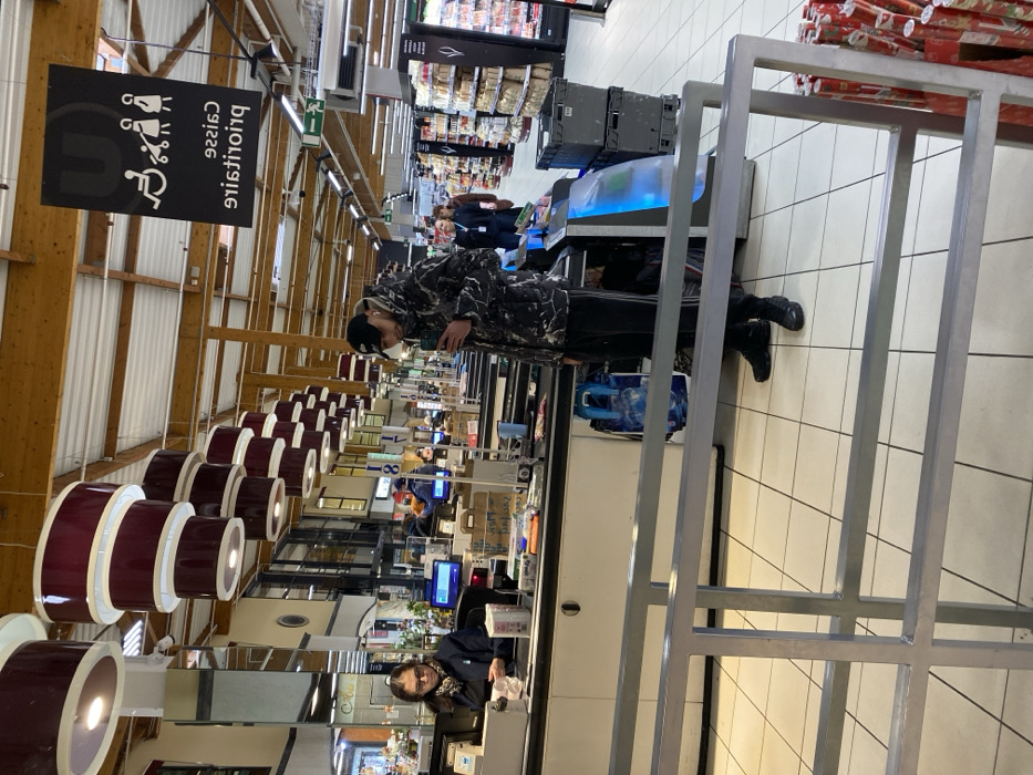 The image depicts the interior of a supermarket checkout area, identified as a "U" brand store (e.g., Super U, Hyper U) by overhead signage, located in Hoenheim, France.
In the foreground-middle, a male customer stands facing slightly right, looking down at a smartphone held in his right hand. He wears a black baseball cap, a grey and white marble-patterned winter jacket, dark athletic pants with two white stripes on the outer seams, and black sneakers. A blue shopping bag rests on the floor near his feet.
To the left, a female cashier is seated at a checkout counter. She wears dark-rimmed glasses and a dark uniform top, with her dark hair pulled back. She appears to be engaged with the cash register or sorting items on the counter. The counter is light-colored and features a conveyor belt, a computer monitor, and various small retail items.
Above the male customer, a black rectangular sign with a white "U" logo is suspended from the ceiling. Below the logo, white pictograms indicate priority access for wheelchair users, pregnant women, and parents with infants. The French text "Caisse prioritaire" (Priority Checkout) is visible beneath the pictograms but appears reversed.
The supermarket ceiling features exposed wooden beams and joists, from which a series of tiered, cylindrical light fixtures with dark red/burgundy exteriors and white interiors are suspended. The floor is covered in large, light-colored square tiles.
In the background, multiple other checkout lanes are visible, along with supermarket aisles stocked with products. Several other individuals, appearing as customers or staff, are discernible further into the store. Grey plastic crates are stacked near an aisle entrance on the right. In the extreme lower right foreground, a stack of red-patterned rolled wrapping paper is visible. A grey metallic barrier or queue rail occupies the lower foreground, extending across the bottom of the frame.