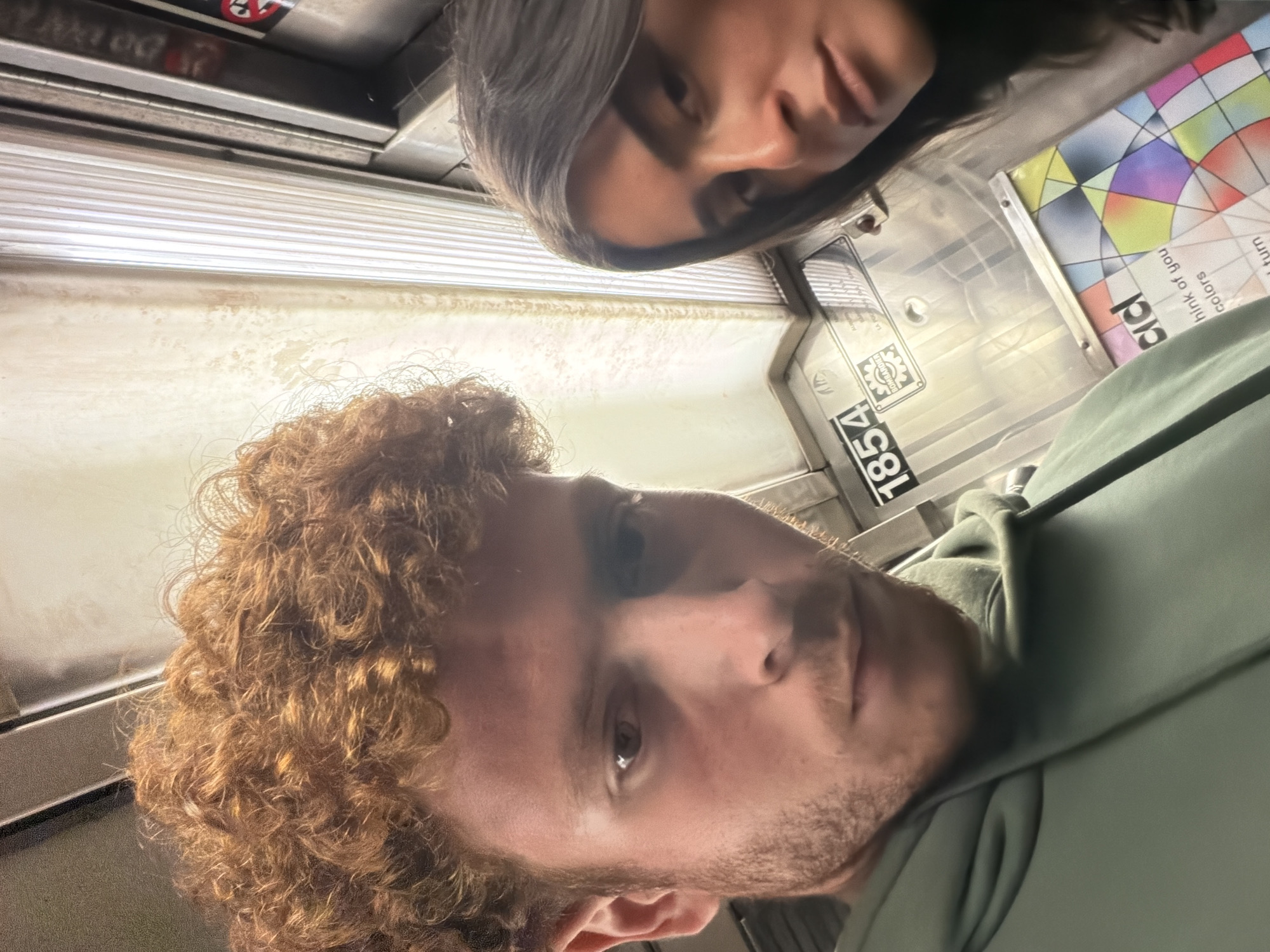 The image captures two individuals, possibly a couple, on a New York City Subway train. The man in the foreground has light skin, reddish-brown curly hair, and a beard. He's wearing a light green hooded sweatshirt. The woman, positioned slightly behind and above him, has dark hair and is looking down. The interior of the subway car is visible in the background, with metal surfaces, an ad poster featuring geometric shapes and text, and a number "1854" displayed, likely part of the train car's identification number. The photo appears to be taken from a low angle.