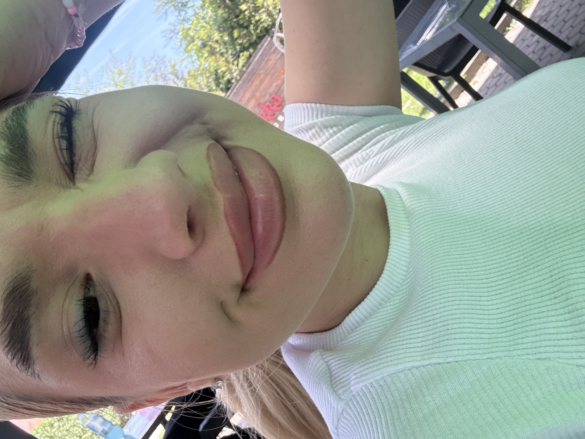 The media file presents a close-up, tilted portrait of a young person, likely female, captured in an outdoor setting in Sofia, Bulgaria, under bright daylight. The subject has light skin, prominent dark eyelashes, and light-colored hair visible at the bottom of the frame. They are wearing a light-colored, possibly white or very light green, ribbed top. A pink and white beaded bracelet is visible on their arm in the upper left.
The person displays a relaxed, joyful, or playful expression, with eyes partially closed and lips slightly parted in a smile, revealing a hint of their tongue. The background is softly blurred but clearly indicates an outdoor environment with green trees and a blue sky, suggesting sunny weather. Out-of-focus outdoor furniture, including dark chairs and a table, are visible, pointing towards a cafe, restaurant, or garden setting. A faint red sign in the background appears to spell "idea." The overall mood is casual and happy.