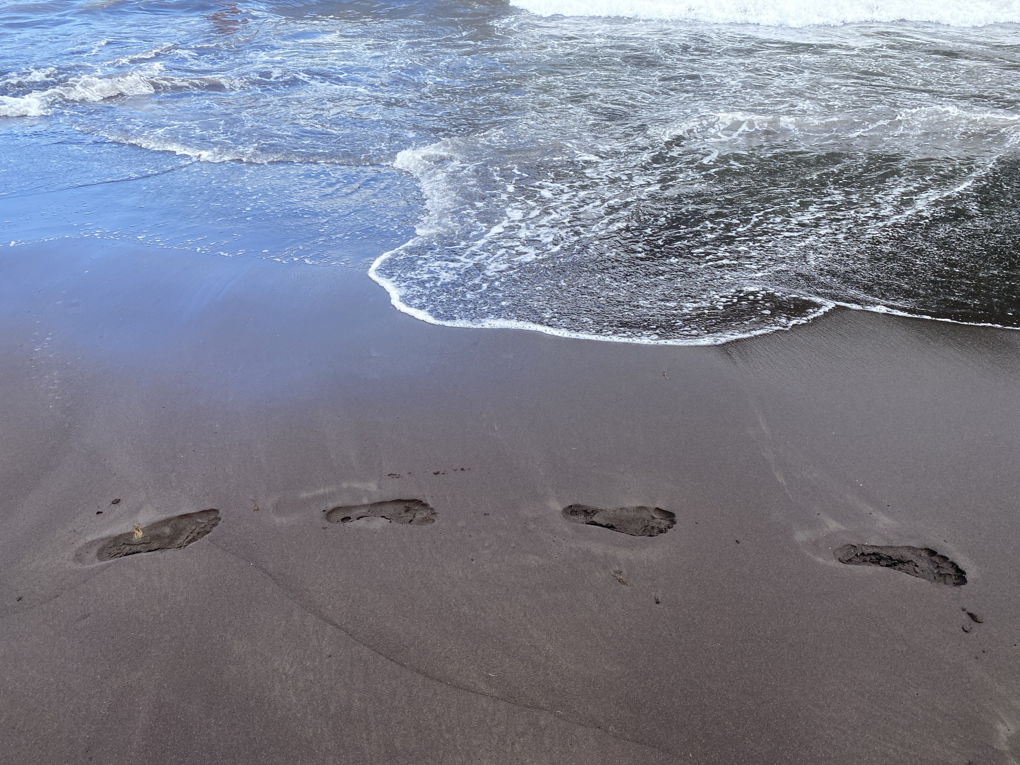The image depicts a beach scene with dark sand and the ocean. Several footprints are visible in the sand, indicating recent human presence. The tide appears to be receding or at a low point, as a line of foam marks the edge of the water where it meets the sand. The water itself is blue and agitated, with small waves and whitecaps. There are no people directly visible in the frame. The location is identified as Porto Moniz, Portugal.