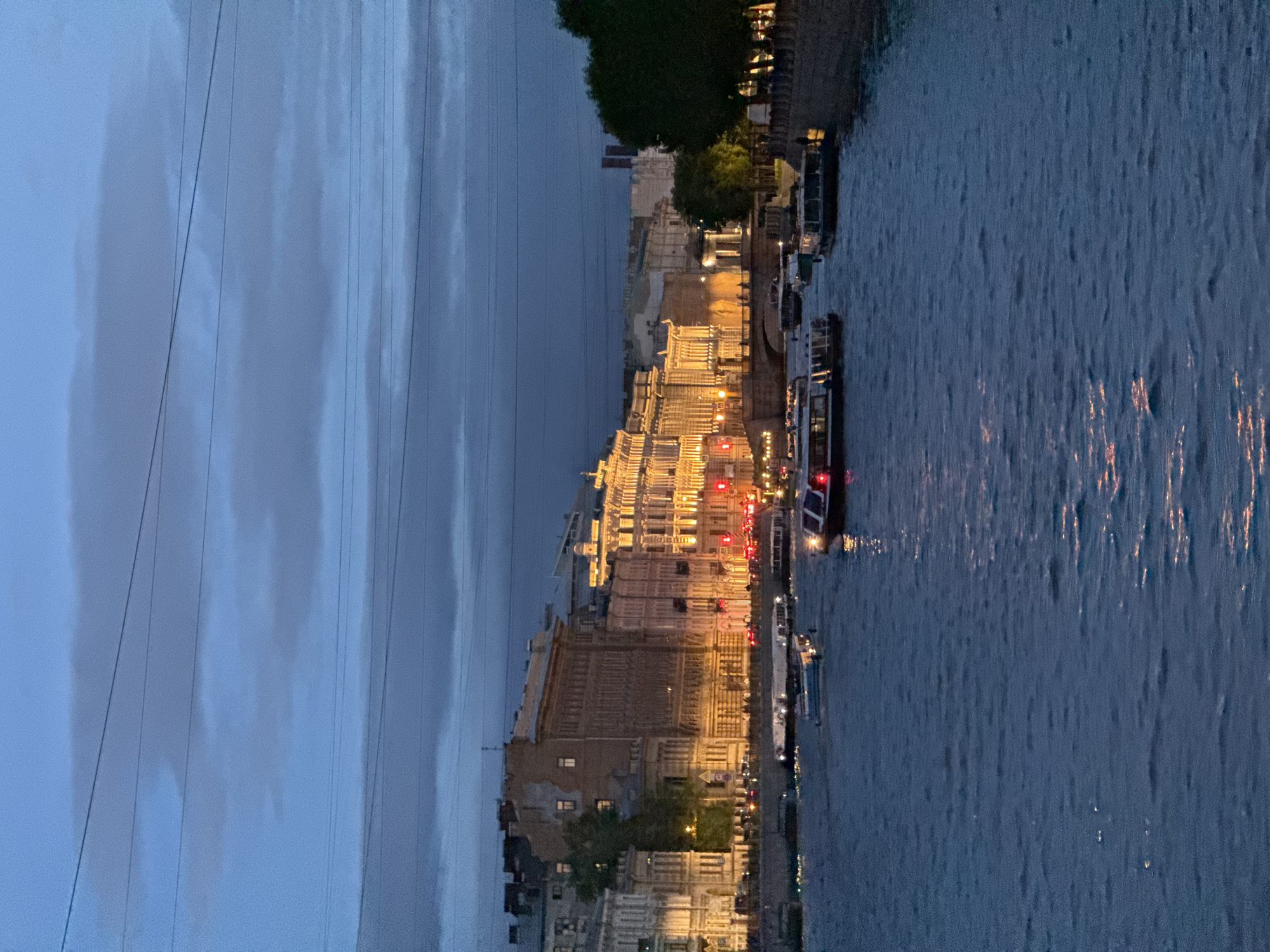 This is a nighttime, eye-level, medium shot of a wide river in Saint Petersburg, Russia. Several boats are on the river, some appearing to be tour boats. The buildings lining the river are illuminated with warm lights, highlighting their architectural details. The sky is a deep blue with dark, cloudy formations and several power lines stretching across it. The water is dark and shows reflections of the city lights.