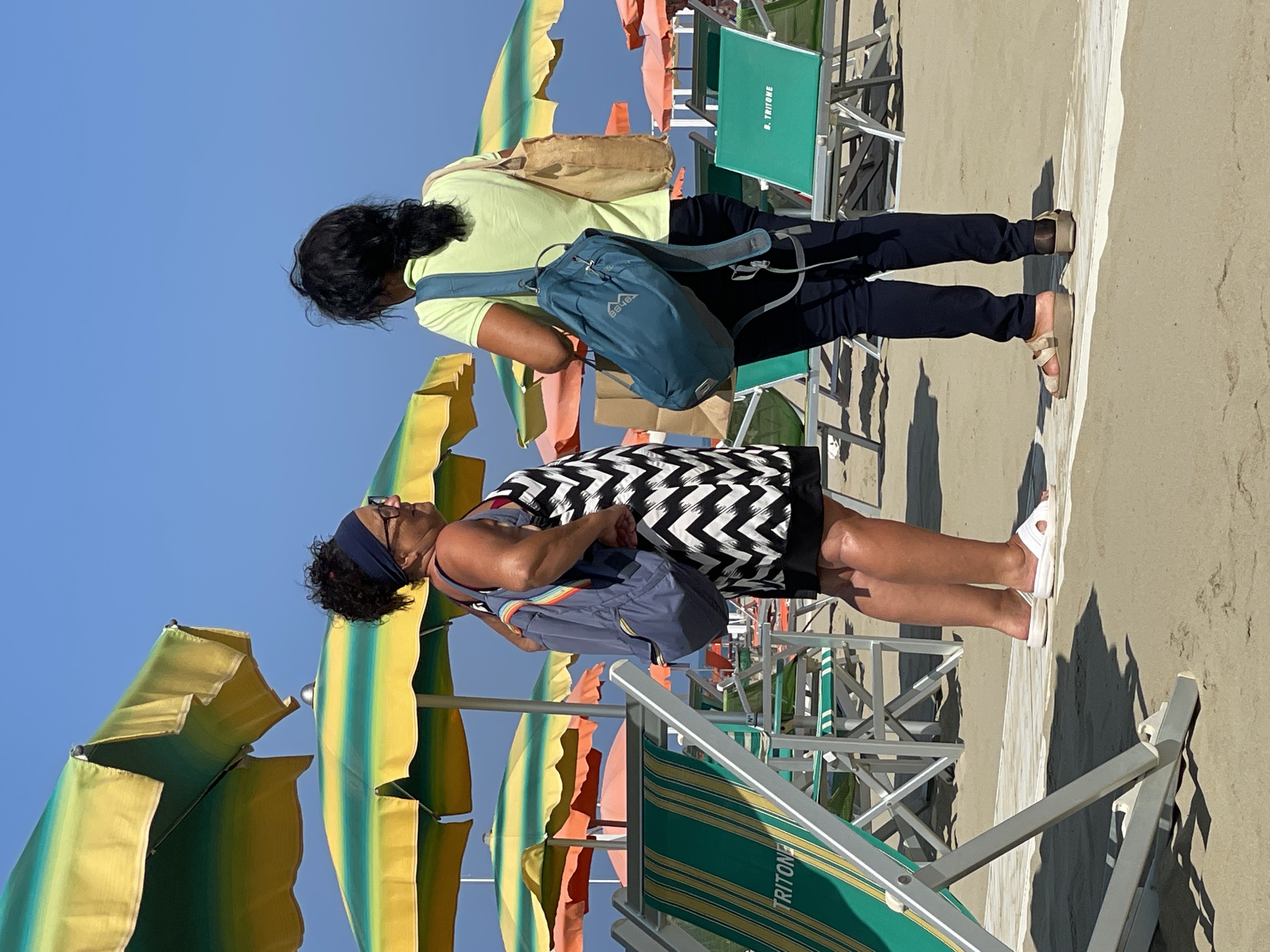 Two women are standing on a sandy beach in Viareggio, Italy, facing each other and engaged in conversation. The woman on the left is wearing a black and white chevron-patterned dress, a dark blue headband, and white slip-on sandals. She has a blue backpack with rainbow-colored straps slung over her left shoulder. The woman on the right is wearing a light green short-sleeved shirt and dark pants, with beige platform sandals. She has a beige tote bag over her right shoulder and a teal backpack on her back.
Behind them, rows of green and white striped beach chairs are set up, many covered by colorful striped umbrellas in shades of yellow, green, and orange. The sky is clear and blue, indicating a sunny day. The sand is light brown, and a white path runs horizontally across the foreground, separating the chairs from the water's edge, though the water itself is not visible. The chairs and umbrellas suggest a well-maintained beach resort.