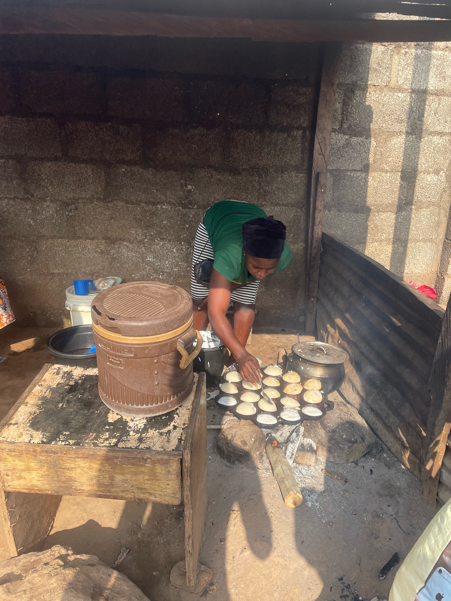In Kurunduma, Nigeria, a woman is cooking small, round pancakes in a pan over an open fire. She is wearing a green shirt and a black headscarf. The pancakes are arranged in a grid in the pan. Next to the pan is a pot with a lid, and behind that are several buckets and containers. The cooking area appears to be outdoors, with a corrugated metal fence to the right and a brick wall to the back. The ground is dirt. Sunlight casts long shadows across the scene.