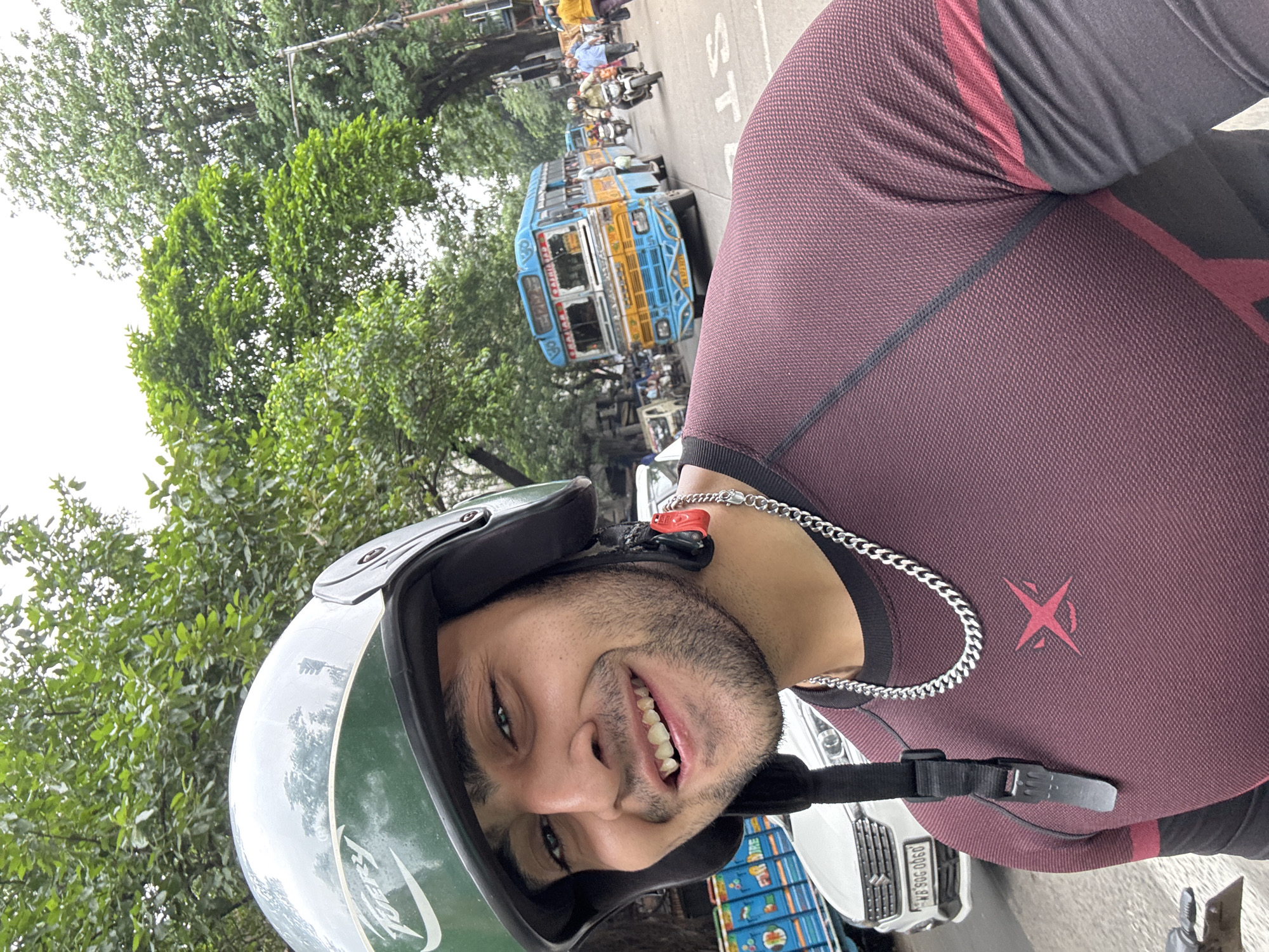 A close-up, selfie-style shot captures a young man wearing a green and silver motorcycle helmet and a maroon long-sleeved shirt with black accents. He is smiling and looking directly at the camera. His face is well-lit, revealing a light beard and a silver chain around his neck. The background is a busy city street in Kolkata, India, featuring a blue and yellow bus, a white car, and other vehicles. Lush green trees line the street, indicating a possibly overcast or cloudy day. The overall impression is of someone enjoying a ride through the city.