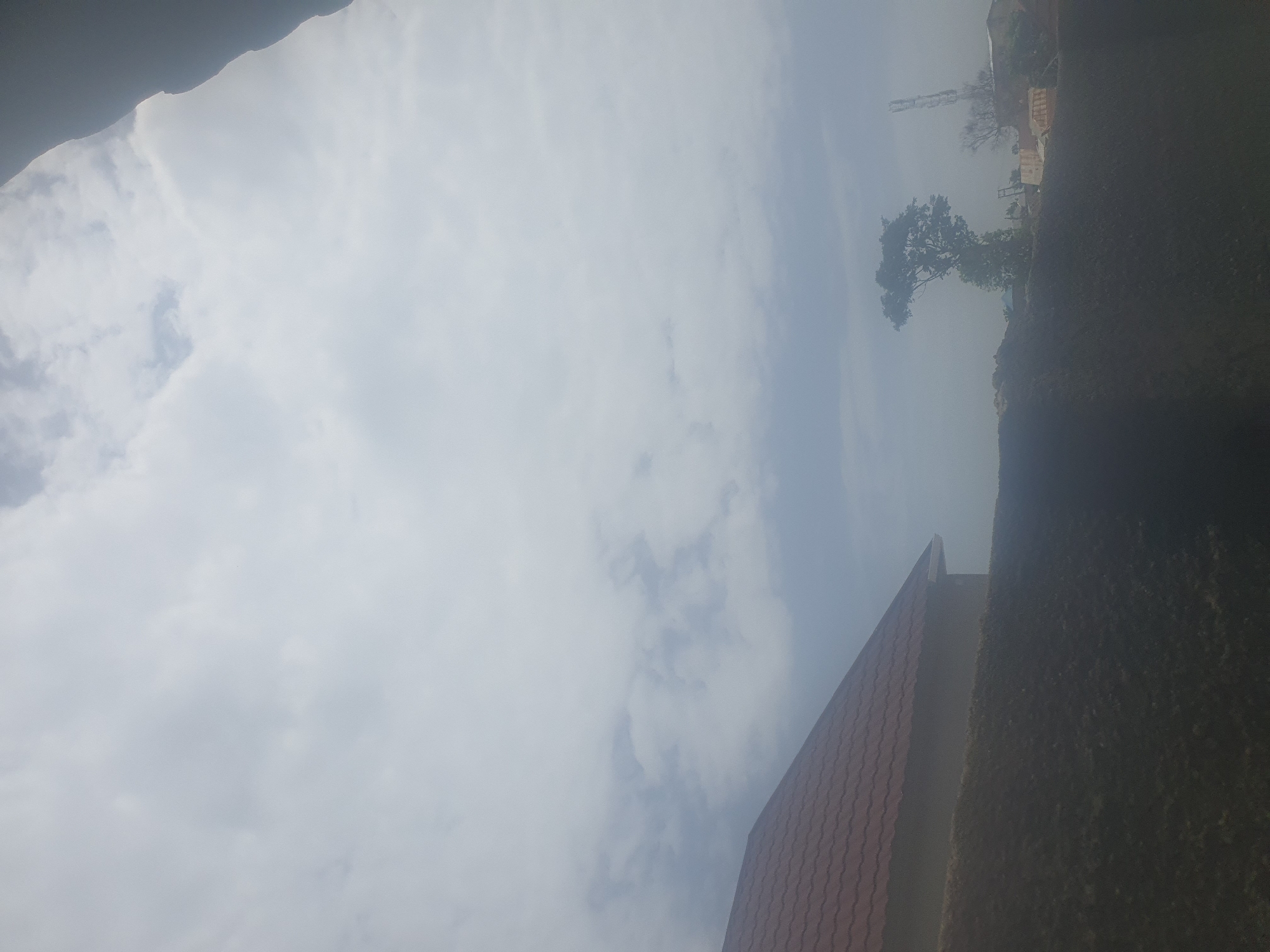 The image shows a cloudy sky over a residential area. On the left, a house with a red tiled roof is visible, partially obscured by a dark, grassy mound in the foreground. In the center-right, a solitary tree stands against the horizon, with a telecommunications tower visible behind it. Some structures and possibly vehicles are present near the tower, suggesting a developing or construction area. The overall impression is of an overcast day in a semi-urban or suburban environment. The location is identified as Jos, Nigeria.