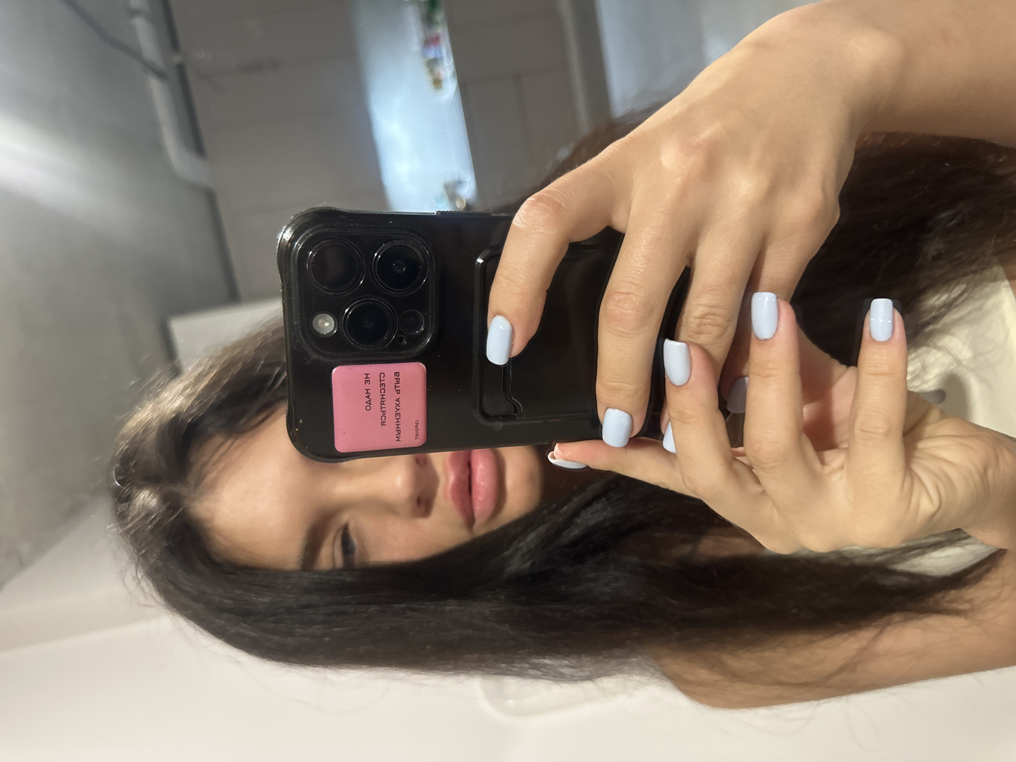 A young woman takes a mirror selfie with a black smartphone. Her dark hair frames her face, which is partially obscured by the phone held in her hands. Her hands are adorned with light blue nail polish on short, square nails. The smartphone case is black and features a prominent multi-lens camera system on the back. A rectangular pink sticker is attached to the phone case, displaying Cyrillic text that is inverted and upside down due to the mirror reflection. The visible text includes characters such as "ОДАН ЭЙ НЕ ЙОТЯА ЙИТЯЭ ЧЛЕЧШЛРCП". Her full lips are visible beneath the phone. The background is a blurry indoor setting with light-colored, reflective surfaces. The overall scene suggests a casual, personal moment captured indoors, possibly in Khimki, Russia.
