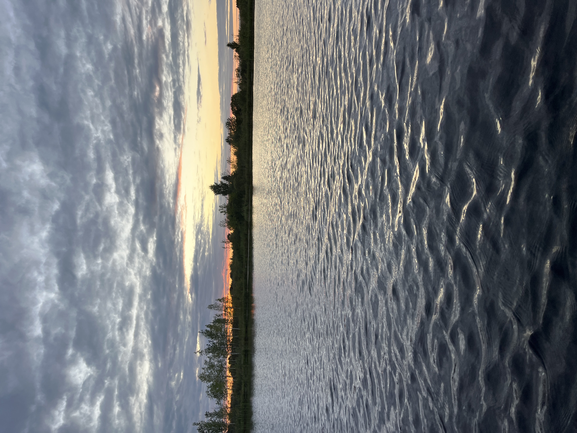 The image depicts a body of water in Навасёлкаўскі сельскі Савет, Belarus, at sunset. The sky is filled with dramatic, dark grey clouds that are beginning to break, revealing streaks of orange and yellow light near the horizon. A line of trees and vegetation forms a dark silhouette against the colorful sky along the distant shore. The water in the foreground is dark and choppy, with small waves reflecting the ambient light. There are no people or man-made structures visible in the scene.