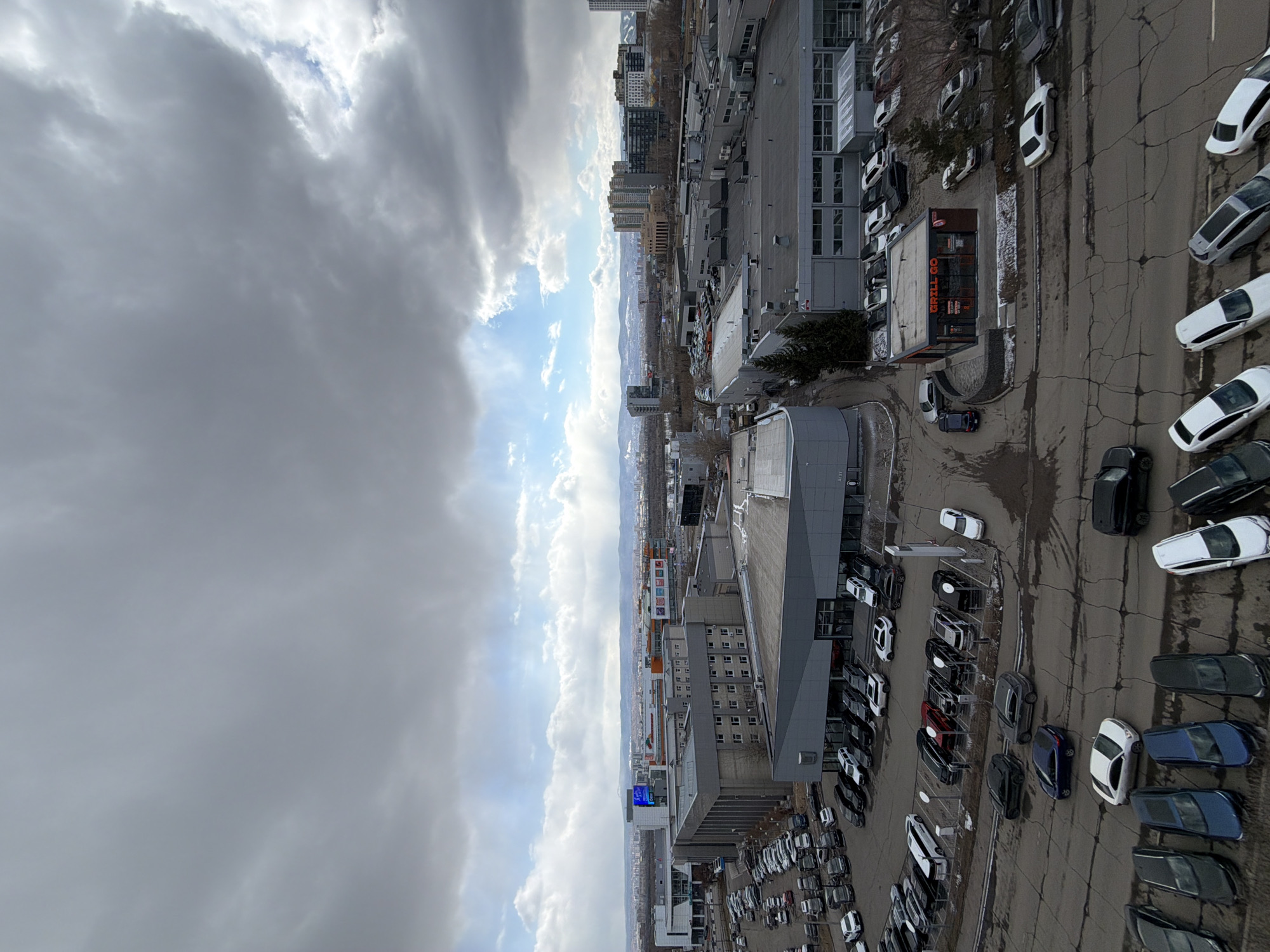 The image depicts an aerial view of an urban landscape, likely in the city of Krasnoyarsk, Russia, given the cityscape and terrain in the background. The foreground is dominated by a commercial area with several large buildings, parking lots filled with cars, and retail establishments. The sky is partially covered with heavy, dark clouds, but there are also patches of blue sky visible. The overall lighting suggests a cloudy day. In the middle ground, there is a visible mountain range in the distance. The city buildings extend into the horizon, interspersed with trees and vegetation. No people are clearly visible in the image. The buildings appear to be a mix of commercial and residential structures, with varying architectural styles. The roads are cracked and worn in places.