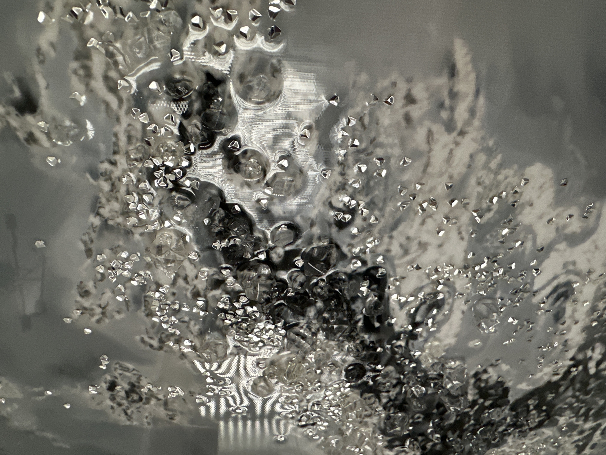 The image shows a close-up, abstract view of small, clear, faceted objects scattered across a textured, gray surface. The objects are irregular in shape and reflect light, giving them a sparkling appearance. Some of the objects appear to be embedded in or floating on a slightly viscous, translucent substance, creating a rippled or bubbly effect. The background is a gradient of gray tones, with subtle patterns that suggest a textured material or light filtering through an uneven surface. There are no people, animals, or identifiable structures visible in the image. The scene appears to be an indoor macro shot, with the location being an unspecified place within an unknown city.