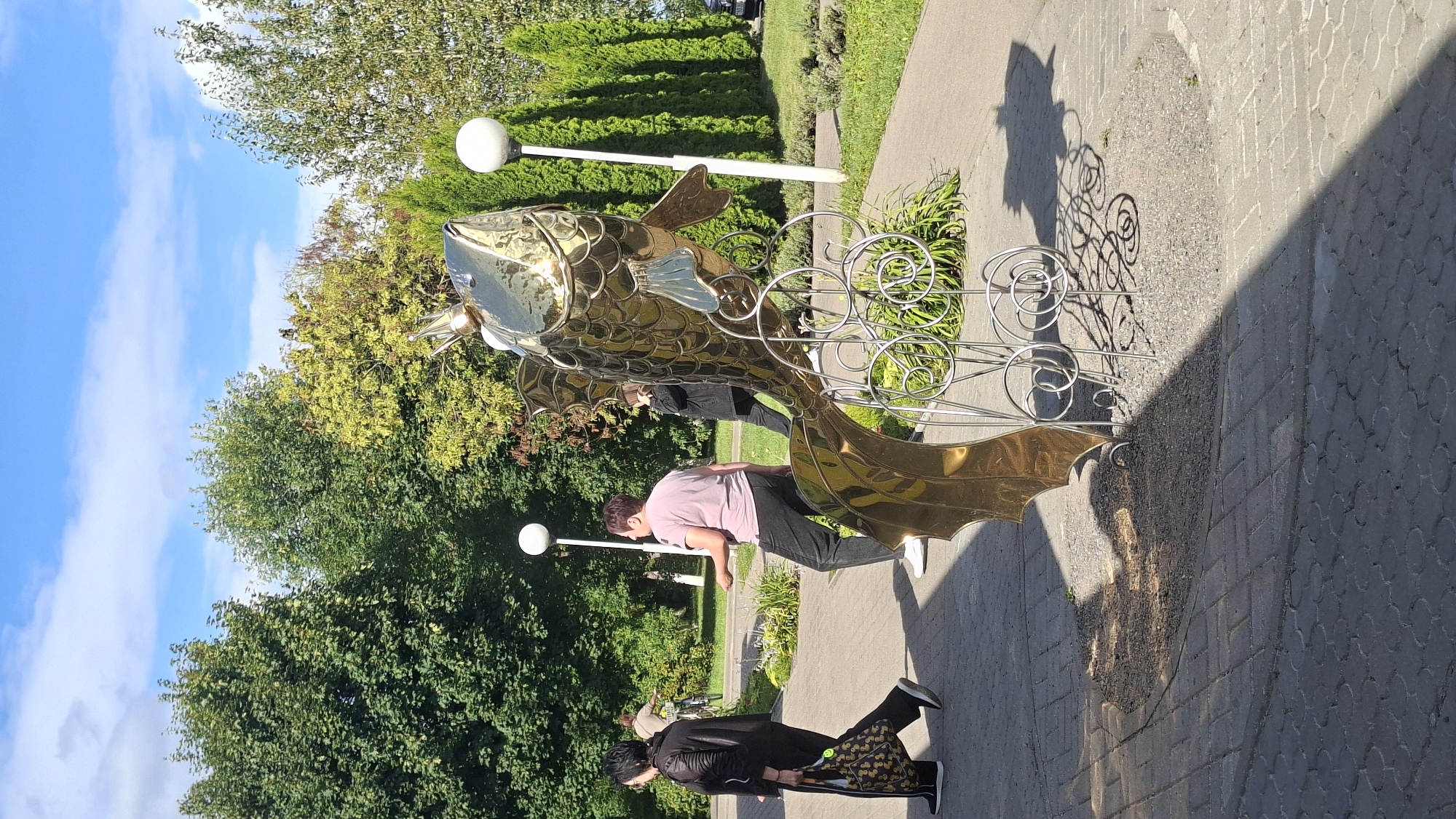 In the city of Myadzyel, Belarus, a large, golden fish sculpture with a crown is positioned on a paved walkway. The sculpture is made of polished metal and features intricate detailing. To the left of the sculpture, two women are walking. The woman in the foreground is wearing a black jacket and pants and carrying a patterned bag. The second woman, slightly behind and to her right, is wearing a pink t-shirt and dark pants. Further back, a person is riding a bicycle. The environment is a park-like setting with lush green trees and manicured hedges. Two white globe-shaped lamps are visible, one to the left of the sculpture and another to its right. The ground is paved with interlocking bricks and gravel. The scene is bathed in sunlight, casting shadows from the sculpture and trees onto the walkway. The sky is blue with scattered white clouds.