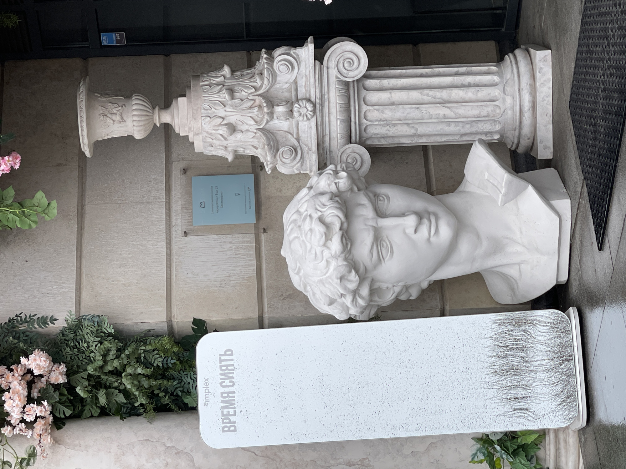 The image shows a display in Saint Petersburg, Russia. There is a white pedestal with the Russian text "ВРЕМЯ СИЯТЬ" (Time to Shine) and the word "simplex". Above the pedestal is a white bust of a male figure, along with a decorative column and vase. These items are set against a wall with green foliage and flowers. The area is likely outdoors, near a building entrance.
