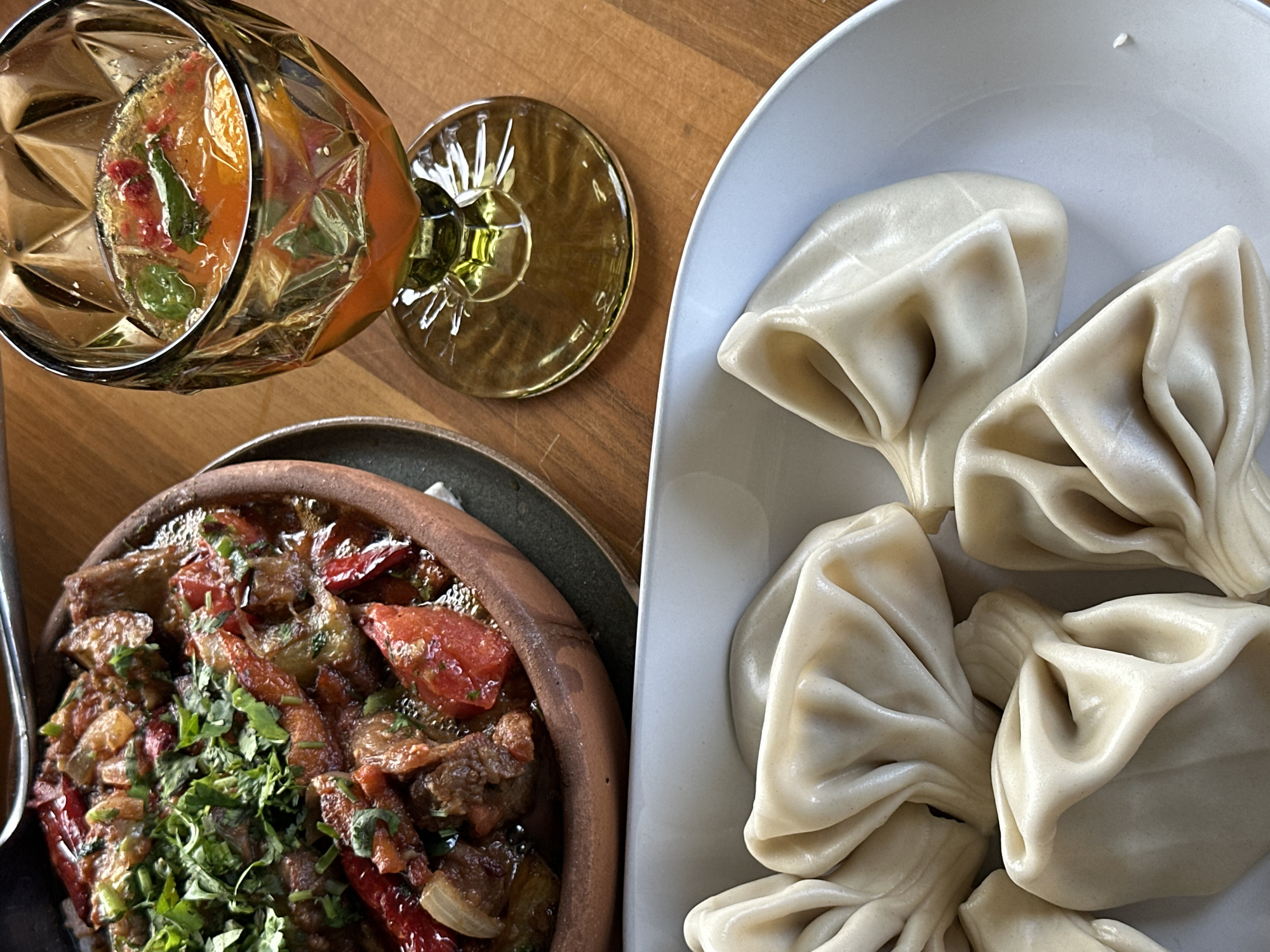 The image captures a dining scene, possibly in Marneuli, Georgia, featuring a selection of Georgian cuisine and a drink. On the right side of the frame, a light blue plate holds four Khinkali, traditional Georgian dumplings characterized by their gathered tops. Adjacent to this, a dark gray earthenware dish contains a meat and vegetable stew, liberally garnished with fresh green herbs. A glass of amber-colored liquid, possibly a cocktail or wine, stands near the stew. The background consists of a wooden table surface. There are no people directly visible in the photograph.