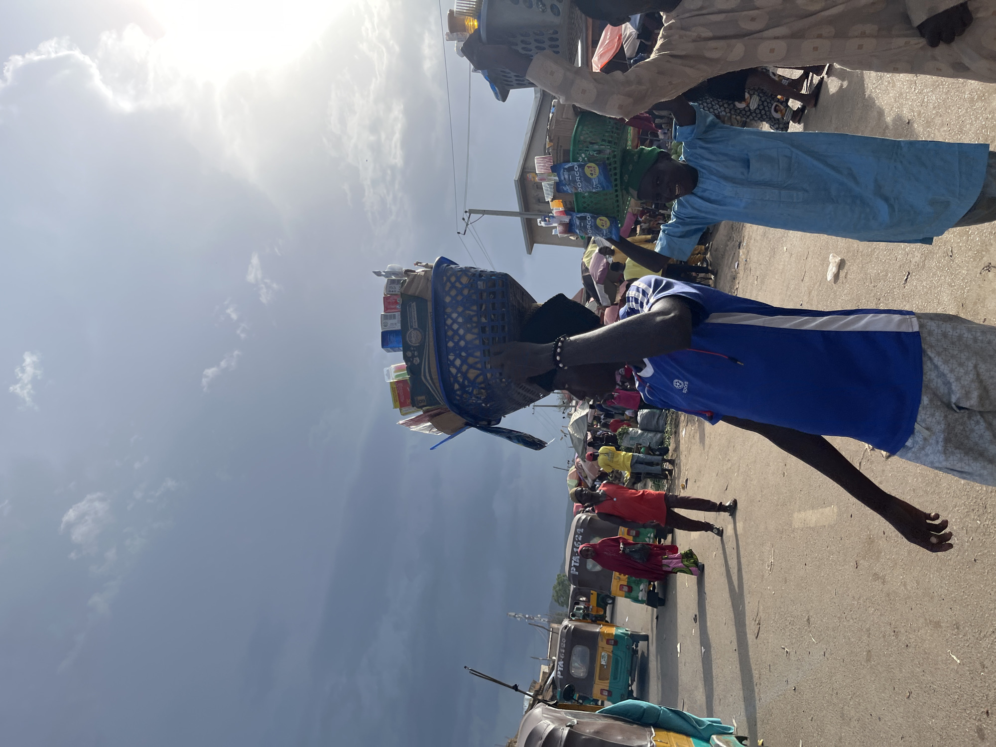 In Rafin Par, Nigeria, a busy market scene unfolds under a partly cloudy sky with the sun peeking through the clouds. Several individuals are walking along a dusty road, some carrying baskets of goods on their heads. In the foreground, a person in a blue t-shirt and patterned shorts walks with their left arm extended. To their right, another person, wearing a light blue traditional outfit and a green head covering, smiles while balancing a green basket on their head. Further to the right, a person in a light-colored garment also carries a basket. In the background, people are milling about in the market, with stalls and awnings visible. Tricycles, a common mode of transport in the region, are also present on the road, with one in the middle ground showing the registration number PTA-6126. The overall atmosphere suggests a vibrant and active marketplace.