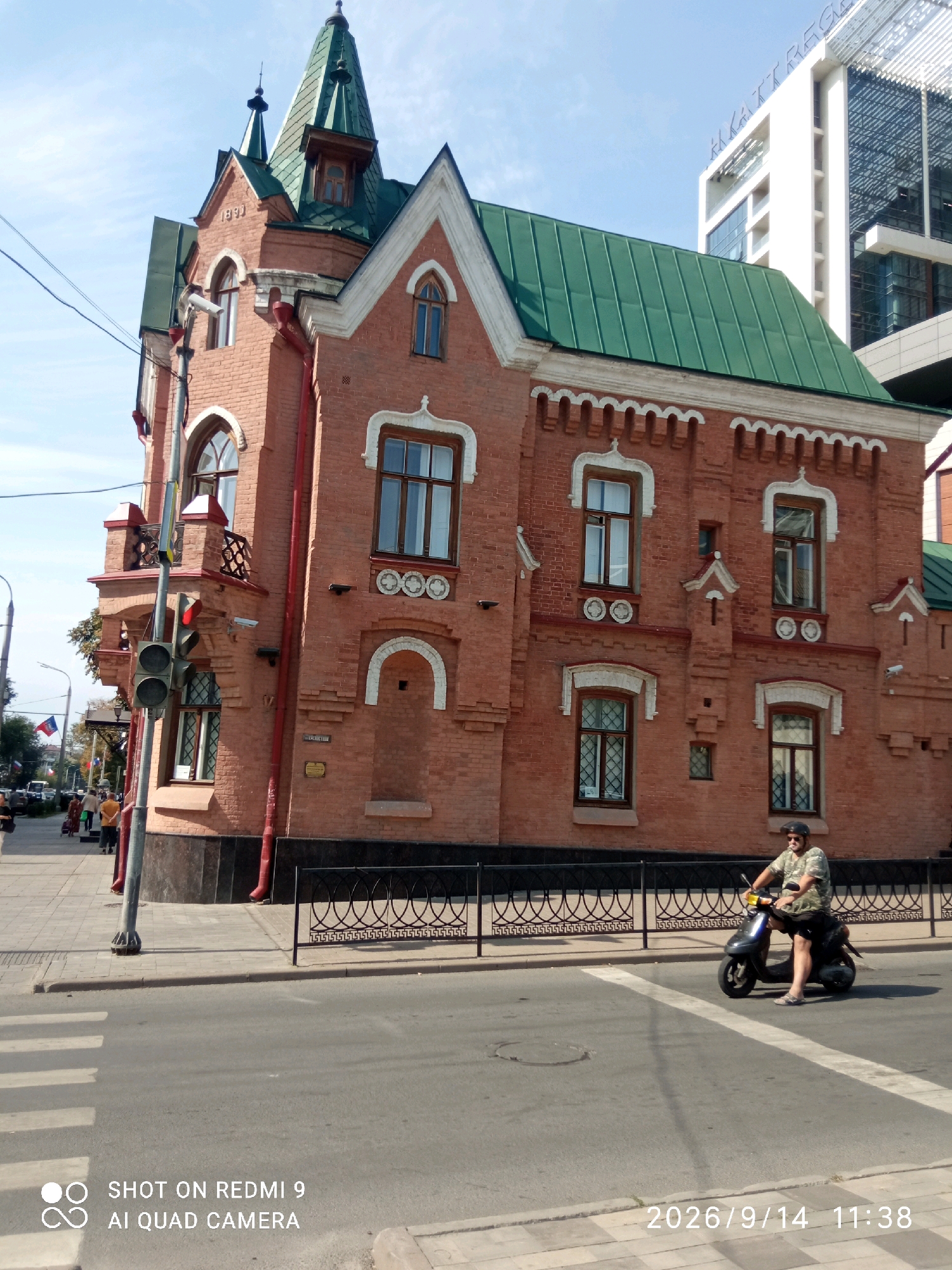 This image depicts a historical red brick building with a green roof, located in Rostov-on-Don, Russia. The building features intricate architectural details, including arched windows and decorative brickwork. A traffic light is visible in the foreground, along with a pedestrian crossing. A person is riding a scooter on the street in front of the building. In the background, a modern white building with glass windows suggests a contrast between old and new architecture in the city. The sky is clear and blue, indicating a sunny day. The presence of flags on poles and people walking on the sidewalk suggests an urban environment. The lower right corner of the image displays a timestamp and camera information: "2026/9/14 11:38" and "SHOT ON REDMI 9 AI QUAD CAMERA".