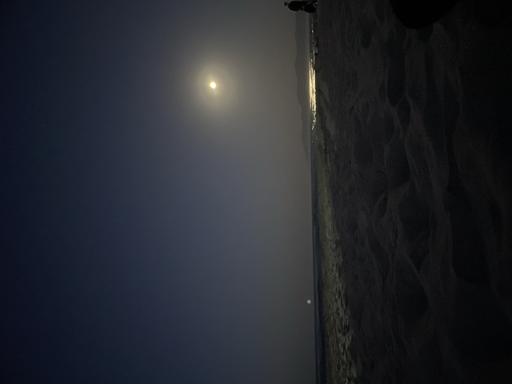 The image depicts a nighttime beach scene. The sky is a deep navy blue, transitioning to a lighter, hazy blue near the horizon. A bright, full moon is visible in the upper right quadrant of the sky, casting a strong glow. Below the moon, the water reflects the moonlight, creating a shimmering path across the sea. The shoreline is composed of sand dunes, which are dark and textured due to the low light. In the distance, along the horizon, a silhouette of mountains can be seen. To the right of the frame, a solitary figure of a person is standing on the beach, facing away from the camera, with another dark object partially visible to their right. The sea appears calm, with gentle waves lapping at the shore. There is a faint, greenish light in the distance over the water, to the left of the frame, possibly another light source or atmospheric phenomenon. The overall impression is one of a quiet, serene night on a beach.