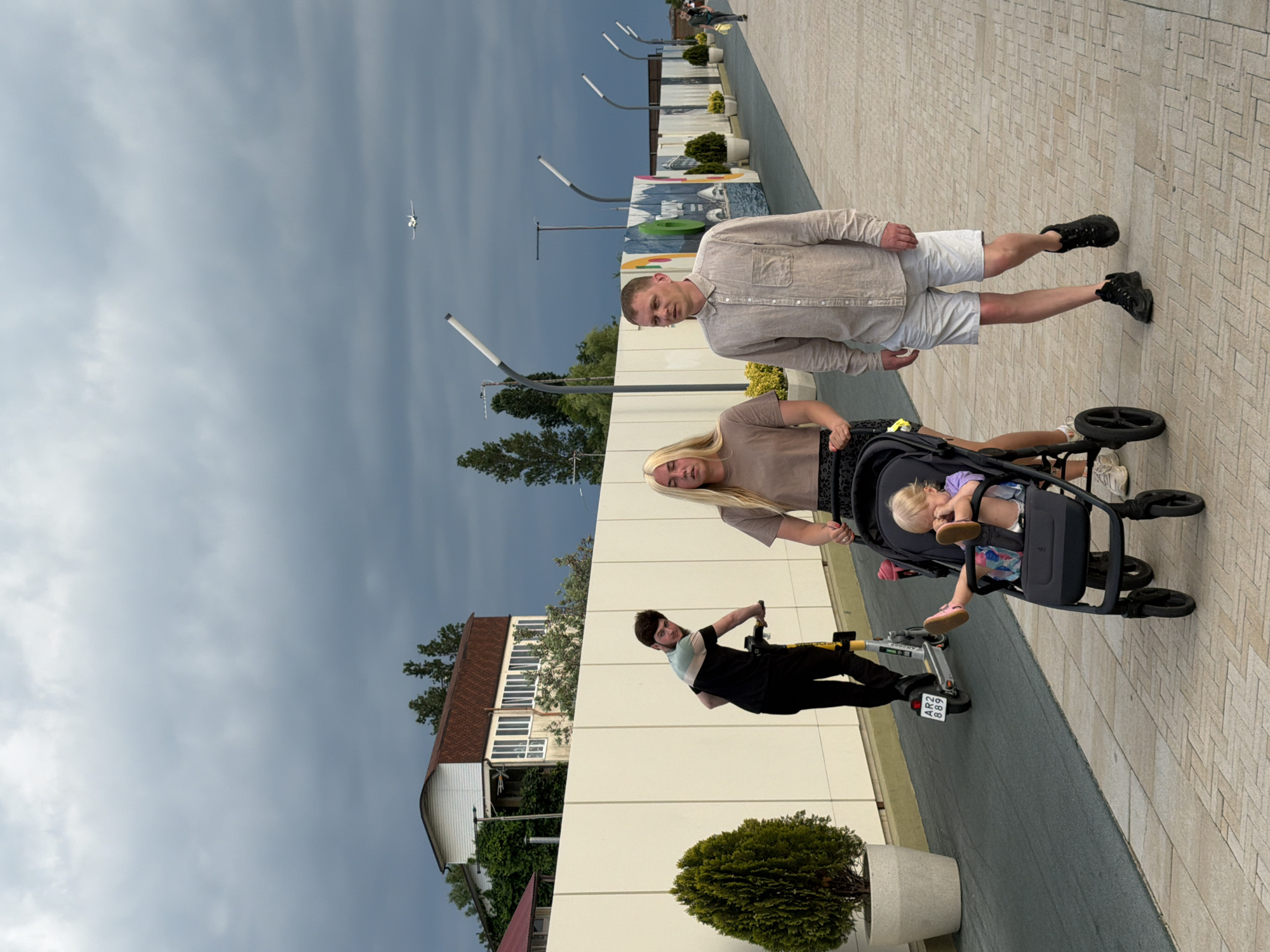 This media file depicts a family scene in Sochi, Russia. The primary subjects are a man, a woman, and a toddler in a stroller, along with another person on an electric scooter. The man is wearing a light-colored linen shirt and white shorts, with black sneakers. The woman has blonde hair and is wearing a tan t-shirt. The toddler, also with blonde hair, is sitting in a dark-colored stroller, dressed in a purple top and patterned bottoms. The person on the scooter is wearing a black and teal striped t-shirt and black pants.
The setting appears to be an outdoor promenade or walkway with a tiled surface. In the background, there is a modern building with a white facade and a red-tiled roof, alongside some greenery and trees. A decorative installation with various colorful elements and lampposts is also visible. The sky is overcast and gray, suggesting a cloudy day. A small airplane can be seen in the sky. The overall mood seems casual and familial, with the individuals engaged in everyday activities. There is some visible text on the license plate of the electric scooter, which appears to be "AR2 889". The time of day is not explicitly indicated, but the lighting suggests it could be afternoon.