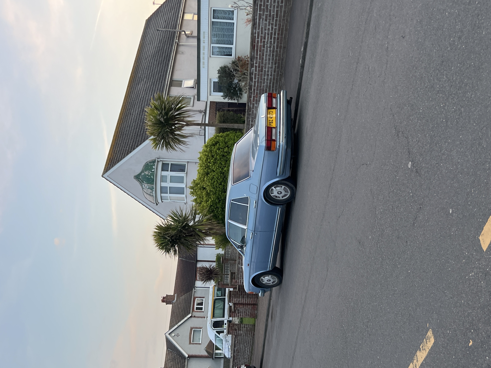 The image shows a light blue vintage Rolls Royce automobile parked on a grey asphalt street in a residential area of Gorleston-on-Sea, United Kingdom. The vehicle is positioned parallel to the curb. Behind the car are a mix of brick and white rendered houses, some with visible palm trees in the front yards. The sky is overcast and light blue in color. Yellow painted road markings are visible on the street. The license plate on the car reads "PBZ 4973". No people are visible in the frame.