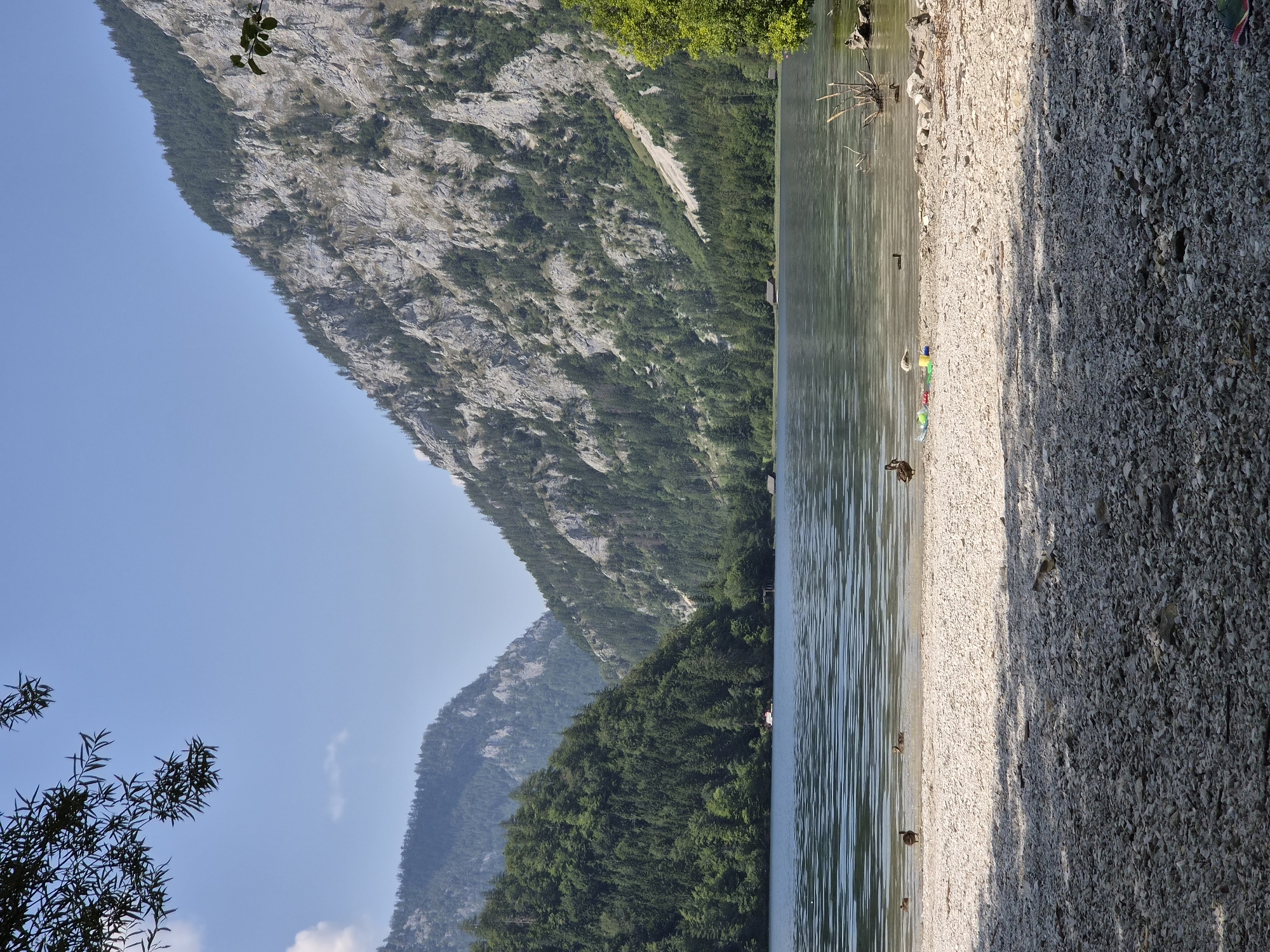 The image depicts a serene lake nestled between towering, tree-covered mountains. In the foreground, a pebble beach slopes gently into the clear, rippling water. Several ducks are visible, some on the shore and others swimming in the lake. Scattered on the beach are a few plastic toys and bottles, suggesting recent human activity.
Further back, across the lake, small wooden boathouses dot the shoreline, interspersed with dense green forestation that climbs the lower slopes of the mountains. The larger mountains rise dramatically behind the treeline, their rocky faces etched with shadows and sunlight. The sky is a clear, bright blue with a few wisps of white clouds.
The scene appears to be located at the Leopoldsteiner See in Eisenerz, Austria, a well-known natural attraction characterized by its stunning mountain backdrop and tranquil lake waters. The overall impression is one of natural beauty and peaceful recreation.