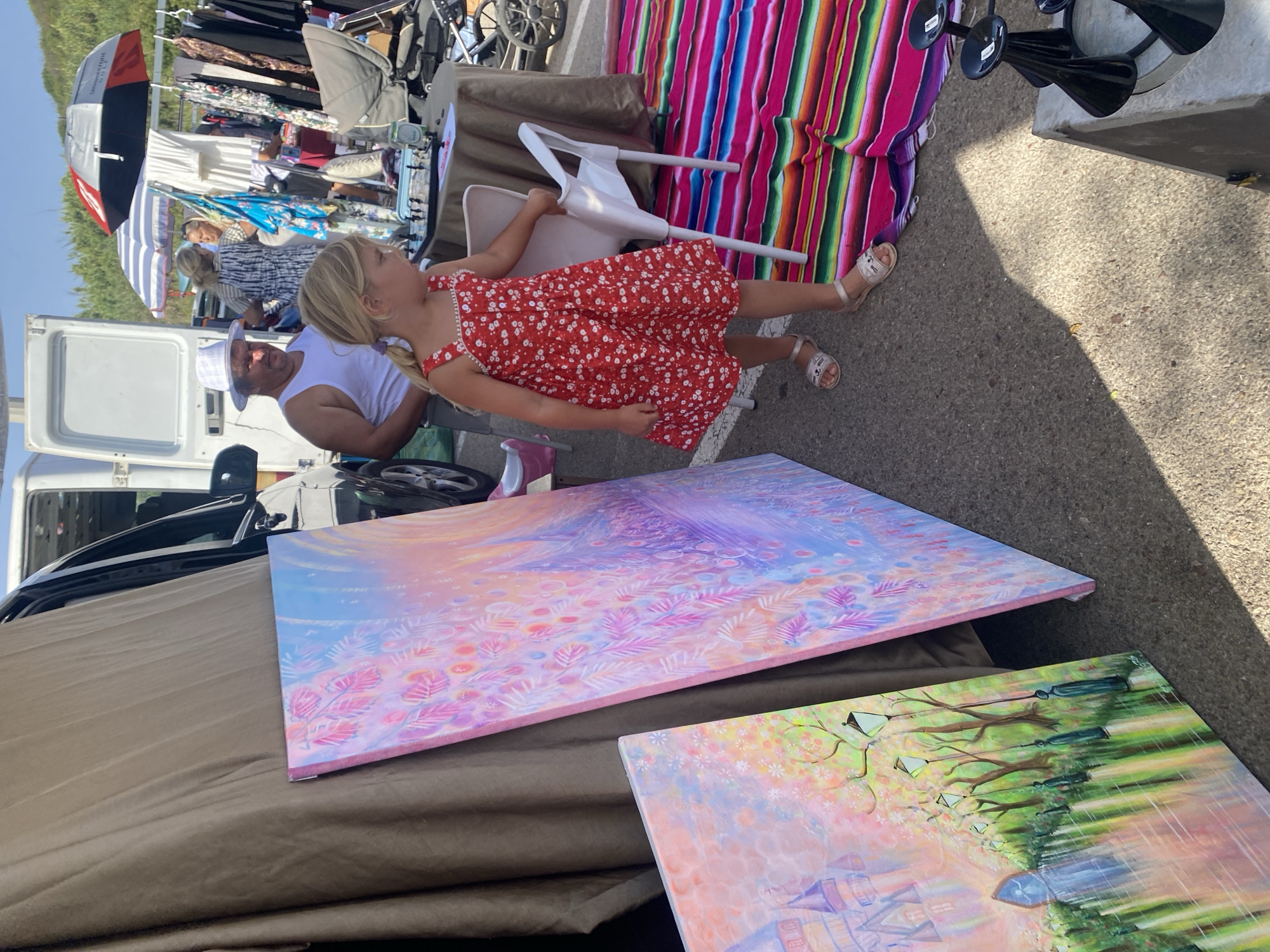The image depicts an outdoor market scene in Manilva, Spain. In the foreground, a young girl with blonde hair in a red floral dress and silver sandals stands near two large paintings. The painting on the left is a landscape with a castle, trees, and lampposts, rendered in soft pastels. The painting on the right is abstract, featuring pink, purple, and blue hues with floral motifs. To the left of the paintings, a portion of a van is visible. In the background, a man wearing a white tank top and a white hat stands near the van. Further back, several other people are browsing stalls with clothing and other items displayed under umbrellas. The ground is paved and lit by sunlight, with shadows cast across the scene. A colorful striped blanket is laid out on the ground near the girl.