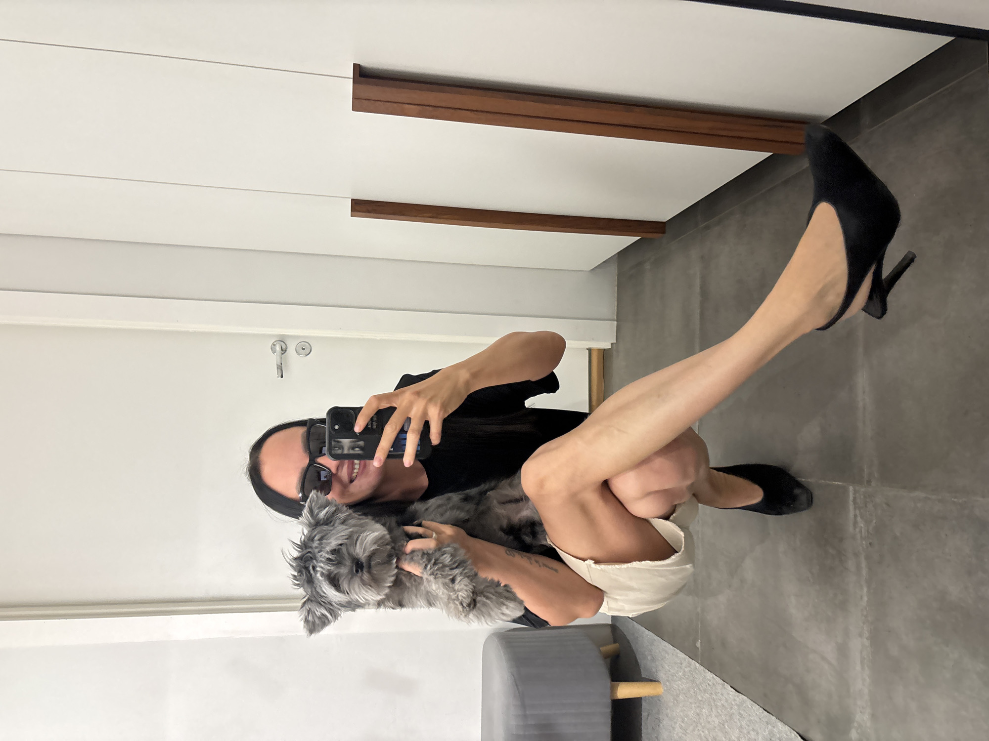 A woman is crouching and taking a selfie in front of a mirror, holding a small gray dog. She is wearing a black top, light-colored shorts, sunglasses, and black high-heeled shoes. Her leg is extended, displaying the heel. The background includes a white wall, wooden trim, a door with a handle, gray tiled flooring, and a gray ottoman. No details indicate location in Chaikovsky, Russia.