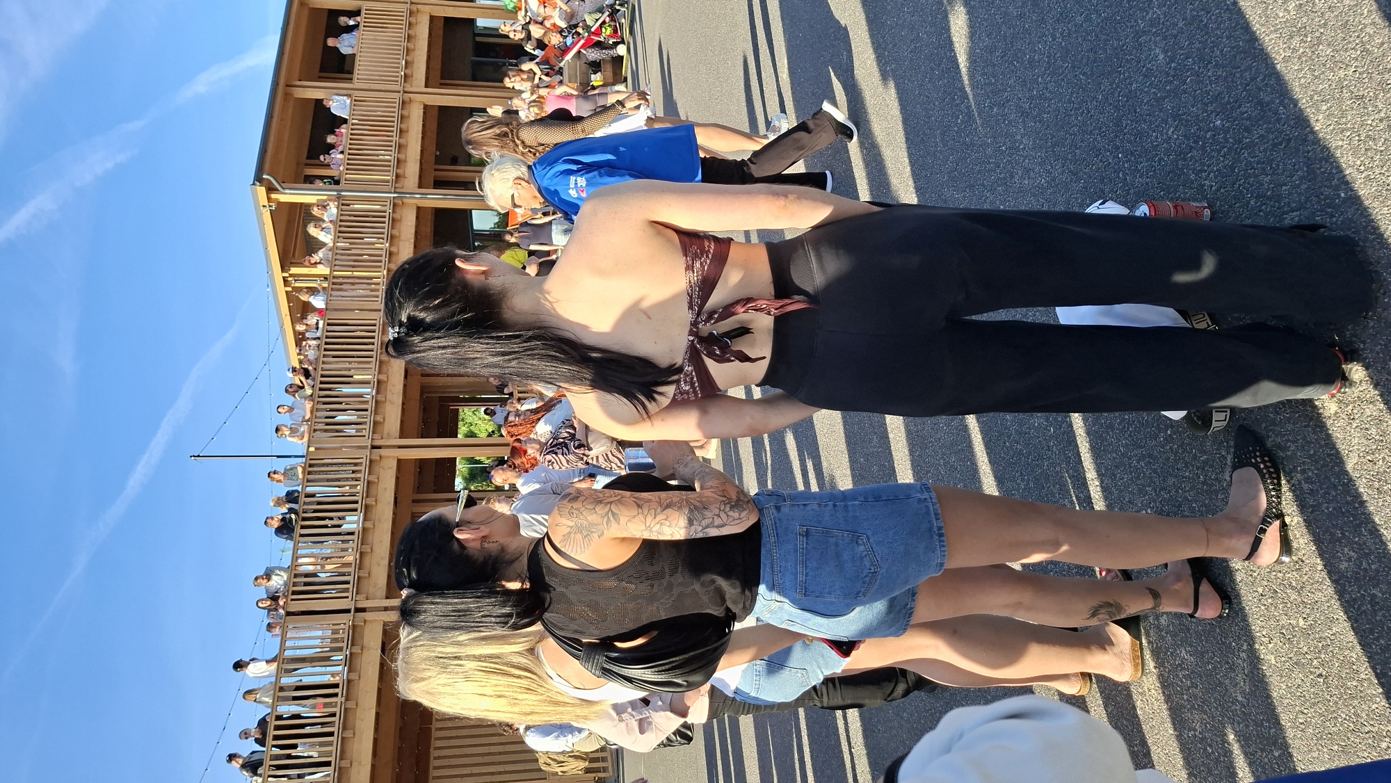 The image captures an outdoor scene with a group of people on a paved surface in front of a modern wooden building with multiple levels and balconies. The sky is clear blue with some faint white contrails. In the foreground, three young women are visible from the back and side. The woman on the left has blonde hair and is wearing a white top and denim shorts. The middle woman has dark hair pulled back in a ponytail, is wearing a black sleeveless top with an intricate pattern and denim shorts. She has extensive tattoos on her arm. The woman on the right has dark hair in a ponytail and is wearing a patterned bandeau top and black wide-leg pants. Several other individuals are in the background, some on the balconies of the building and others mingling on the ground. The building appears to be a public space, possibly a restaurant or event venue, with lighting fixtures visible along the upper railing. The overall atmosphere seems casual and social. The location is identified as Tallinn, Estonia.