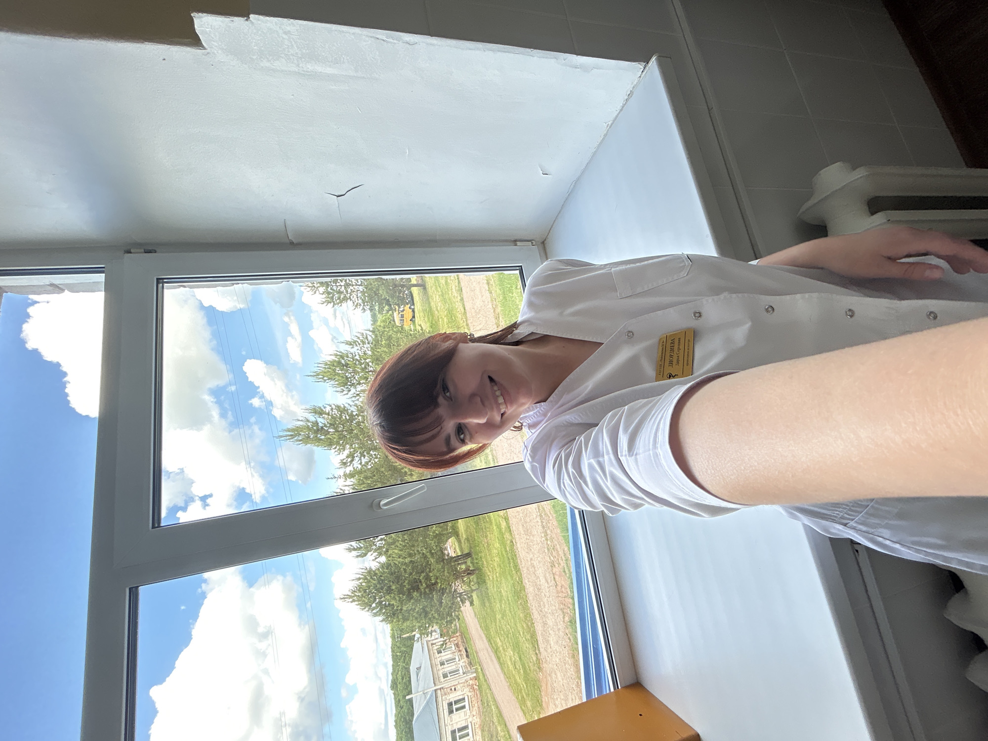 The image shows a person in a white lab coat standing by a window in what appears to be an indoor setting. The background visible through the window suggests a rural or suburban area, with trees, green fields, and buildings. The location is Фоки, Russia. The person is smiling at the camera.