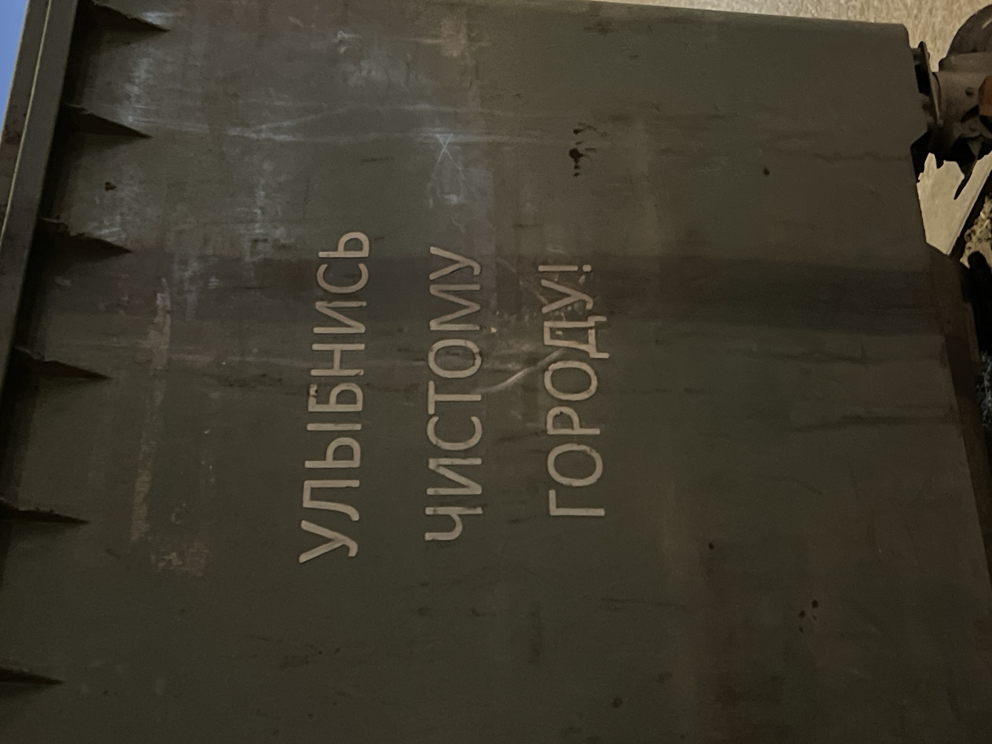 The image depicts the side of a dark green dumpster. Vertically written in Cyrillic script on the dumpster are the words: УЛЫБНИСЬ ЧИСТОМУ ГОРОДУ!, which translates to "Smile to a clean city!". The dumpster appears to be outdoors. The photo was taken in the city of Krasnodar, Russia. The scene appears to be illuminated by artificial lighting. There are no people visible in the frame.