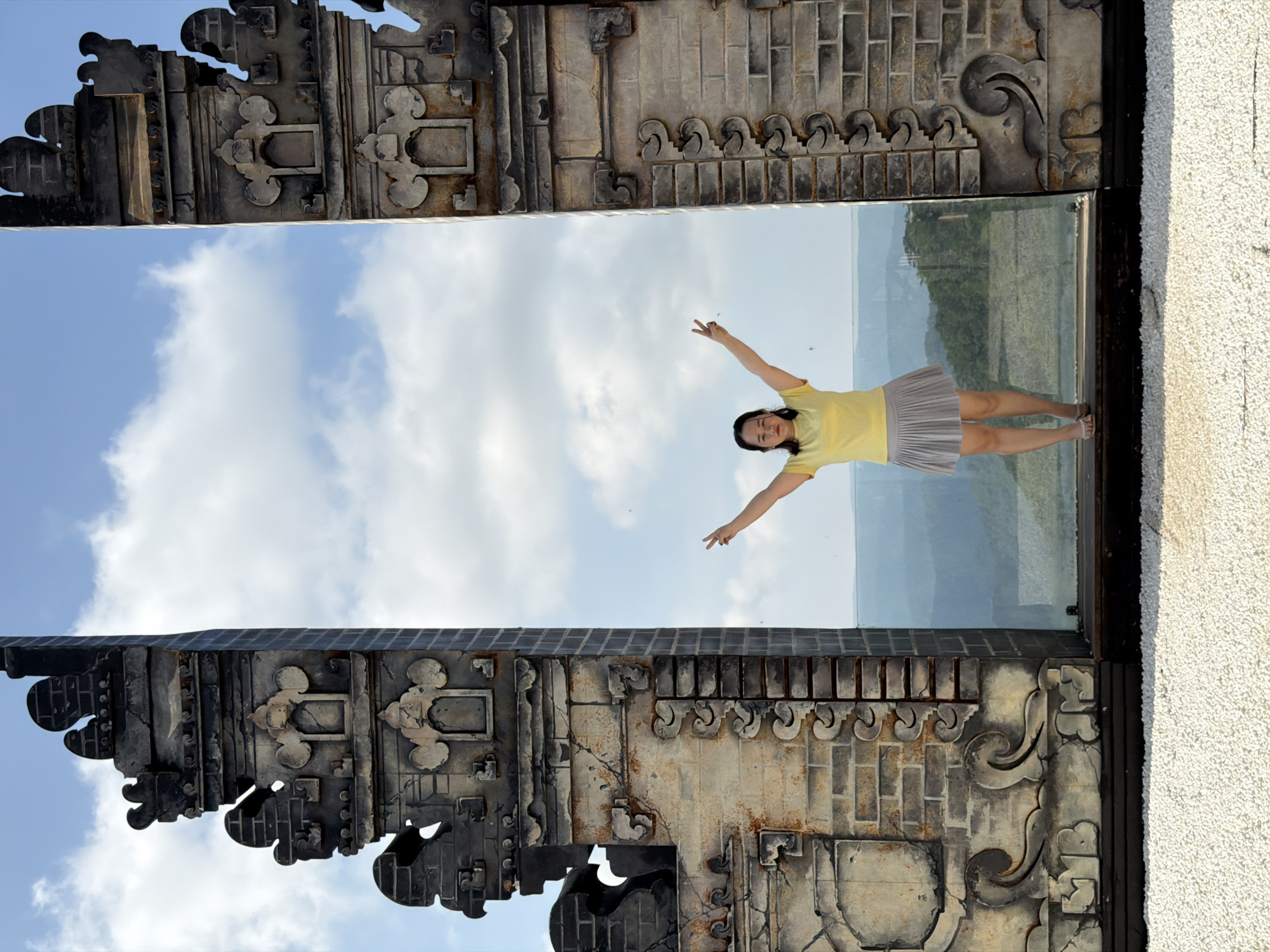 A woman stands between two ornate stone gates, her arms raised in a V-shape, in front of a scenic overlook in Da Lat, Vietnam. The gates are weathered and textured, with intricate carvings depicting floral and cloud motifs. The woman is wearing a yellow t-shirt and a gray pleated skirt. Behind her, a vast landscape unfolds with rolling hills and a hazy sky filled with clouds. The foreground is covered in white gravel. The location is an elevated viewpoint, likely a tourist attraction, offering panoramic views of the surrounding mountainous terrain.