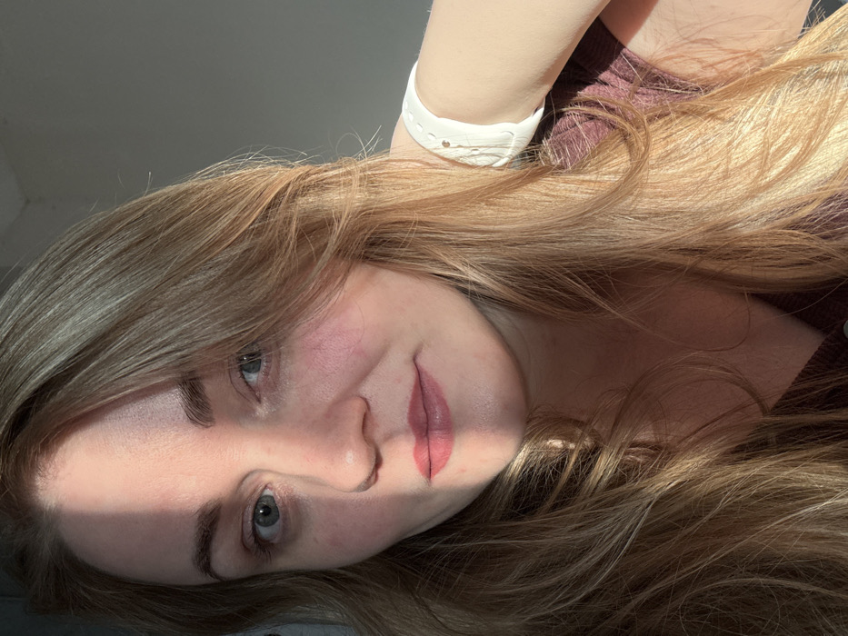 A close-up portrait depicts an adult female indoors, in the city of Chaikovsky, Russia. The individual has fair skin, long, wavy light-brown hair, and light blue eyes, gazing directly forward. Direct sunlight illuminates the right side of her face (viewer's right), creating a sharp shadow line that extends across the bridge of her nose, her left cheek, and the left side of her mouth. Her visible lips are colored with a light reddish tint. A white, textured wristband or watch is visible on her right wrist, positioned against her neck/shoulder area. She is wearing a dark reddish-brown, possibly ribbed or textured, garment with a visible seam or button detail near the neckline. The background consists of a plain, light gray surface on the left (viewer's left) and a darker, undefined area on the right (viewer's right). No other individuals or specific environmental features are discernible.