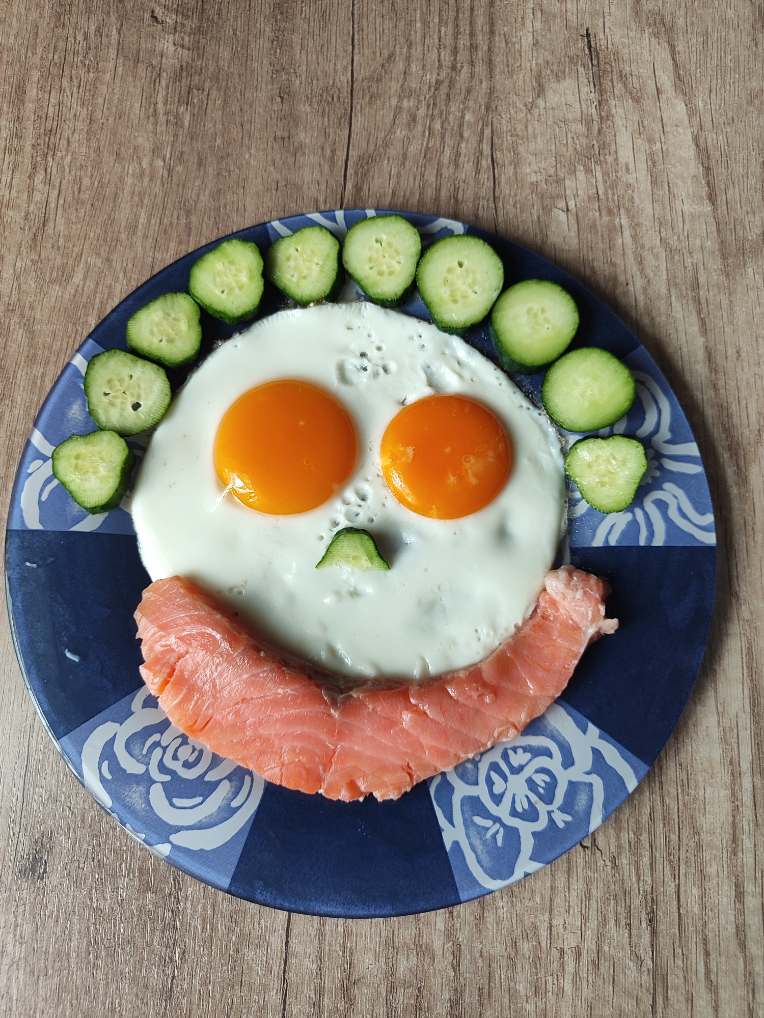 A close-up, overhead shot displays a plate of food arranged to resemble a smiley face. The plate, which has a dark blue rim with a white floral pattern, rests on a wooden table.
The "face" is comprised of:
- Two fried eggs, forming the eyes, with their bright orange yolks prominent against the white of the egg whites.
- A sliver of cucumber positioned between the eggs, serving as a nose.
- A curved slice of smoked salmon laid across the bottom half of the plate, acting as a smiling mouth.
- Slices of cucumber arranged in an arc around the top half of the plate, resembling eyebrows or hair.
The food is centrally positioned on the plate, with a portion of the wooden table visible around it. The lighting appears to be natural, casting a soft illumination on the scene. There is no indication of a specific location in Almaty, Kazakhstan.
