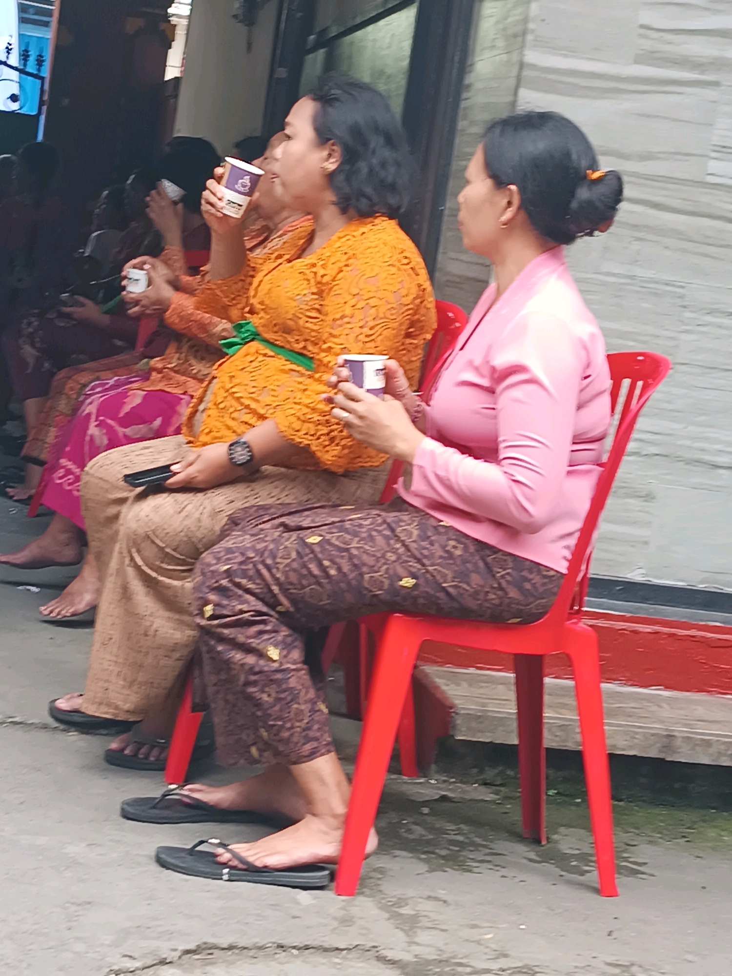 In Tabanan, Indonesia, two women are seated on red plastic chairs. The woman on the left wears an orange lace top and patterned pants, holding a white cup. The woman on the right wears a pink satin top and patterned pants, also holding a white cup. Several other people are visible in the background, some seated, with one person holding a cup. The environment appears to be outdoors, with a tiled wall in the background and a concrete ground. The overall scene suggests a gathering or event in a public space.