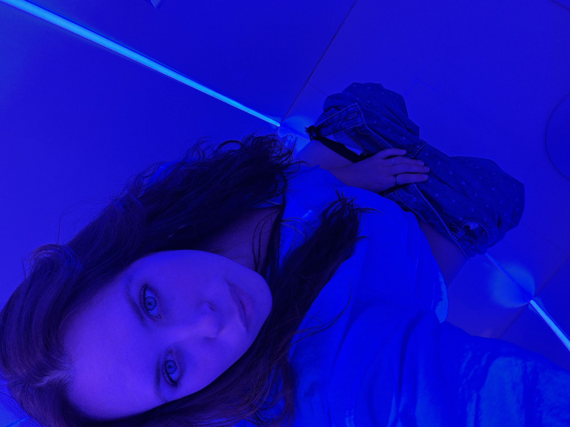 The image captures a young adult female from a high-angle perspective, centered in a brightly illuminated blue environment. She has long, dark hair with a central part and light-colored eyes, which appear intensely blue due to the lighting. Her facial expression is neutral, with her gaze directed straight towards the camera. She is wearing a light blue long-sleeved top. Her left hand, adorned with a ring on the ring finger, is resting on a denim-textured item, possibly a bag or folded garment, held against her lower body. The background consists of smooth, planar surfaces (walls and floor) uniformly bathed in a deep, vibrant blue light. Linear blue LED light strips are visible, integrated into the junctions of the walls and floor, creating a stark, modern aesthetic. A white, curved, semi-circular object is visible on the floor in the bottom right corner. The specific location within Moscow, Russia, cannot be determined from the visual information provided.