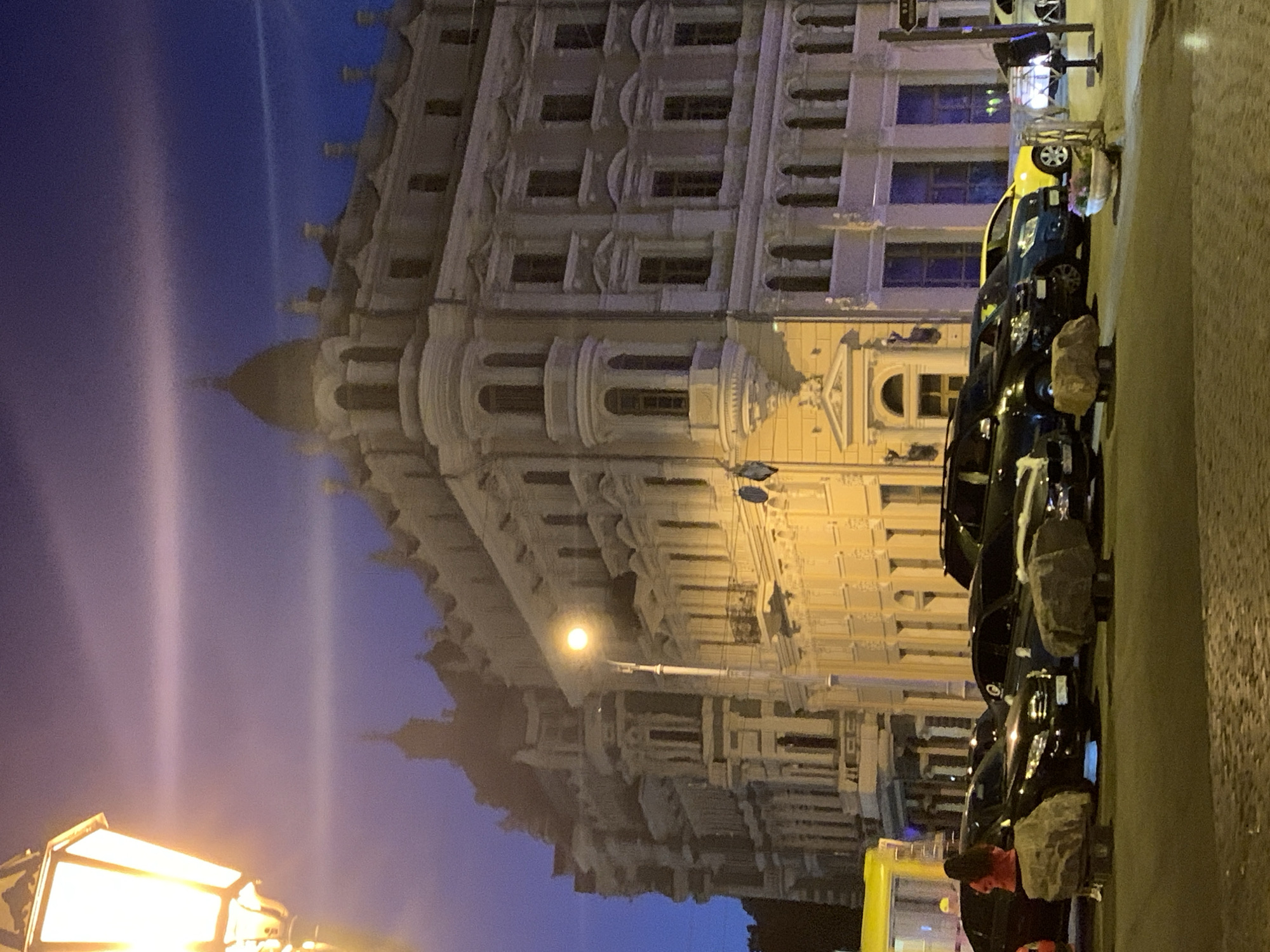 In Odesa, Ukraine, at twilight, a street scene unfolds with a prominent, ornate building as the backdrop. The building, bathed in the warm glow of streetlights, displays classical architectural elements like columns, arched windows, and decorative cornices. In the foreground, a line of parked cars, including a dark SUV and a bright yellow sedan, are visible on the street. A lone figure in a red jacket stands near a parked car, facing away from the camera. Large, rough-hewn stones, likely decorative planters or bollards, are placed in front of the vehicles. The sky above is a deep twilight blue, with a subtle horizontal band of light suggesting a high cloud or atmospheric phenomenon. A street lamp with a warm, illuminating light is positioned on the left side of the frame, casting a soft glow. The overall impression is a quiet urban evening, with the grandeur of the architecture contrasting with the everyday presence of vehicles and a solitary pedestrian.
