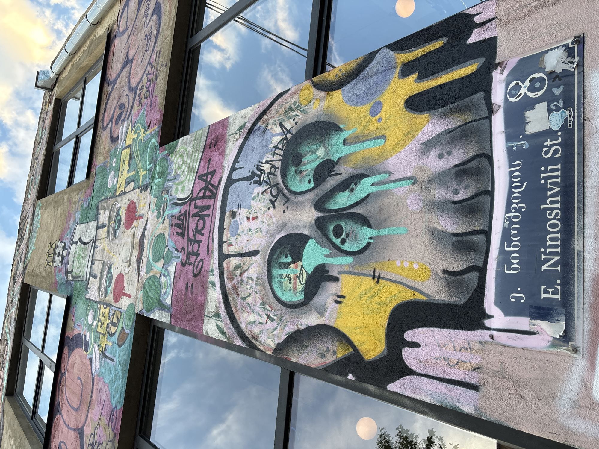 This image shows a close-up, angled view of a building exterior covered in vibrant graffiti. The building itself appears to be modern, with large windows reflecting a cloudy sky. The graffiti is a mix of abstract shapes, characters, and text, rendered in a variety of bright colors including yellow, teal, pink, purple, black, and green.
Dominating the center of the frame is a stylized face with large, circular eyes rendered in teal and black. The face is part of a larger, colorful abstract design. To the right of the graffiti, there is a street sign that is partially obscured by the artwork. The sign is dark blue and has white text. Visible text on the sign appears to be in both English and what looks like Georgian script. The English text reads "E. Ninoshvili St." and above that, "j. 606vıL'ı". The Georgian text reads "ლ. ნინოშვილი". There are also some numbers visible, including "8" and "2". The overall impression is one of urban art and a busy street scene. The lighting suggests it might be daytime, possibly late afternoon given the soft light in the sky reflection.