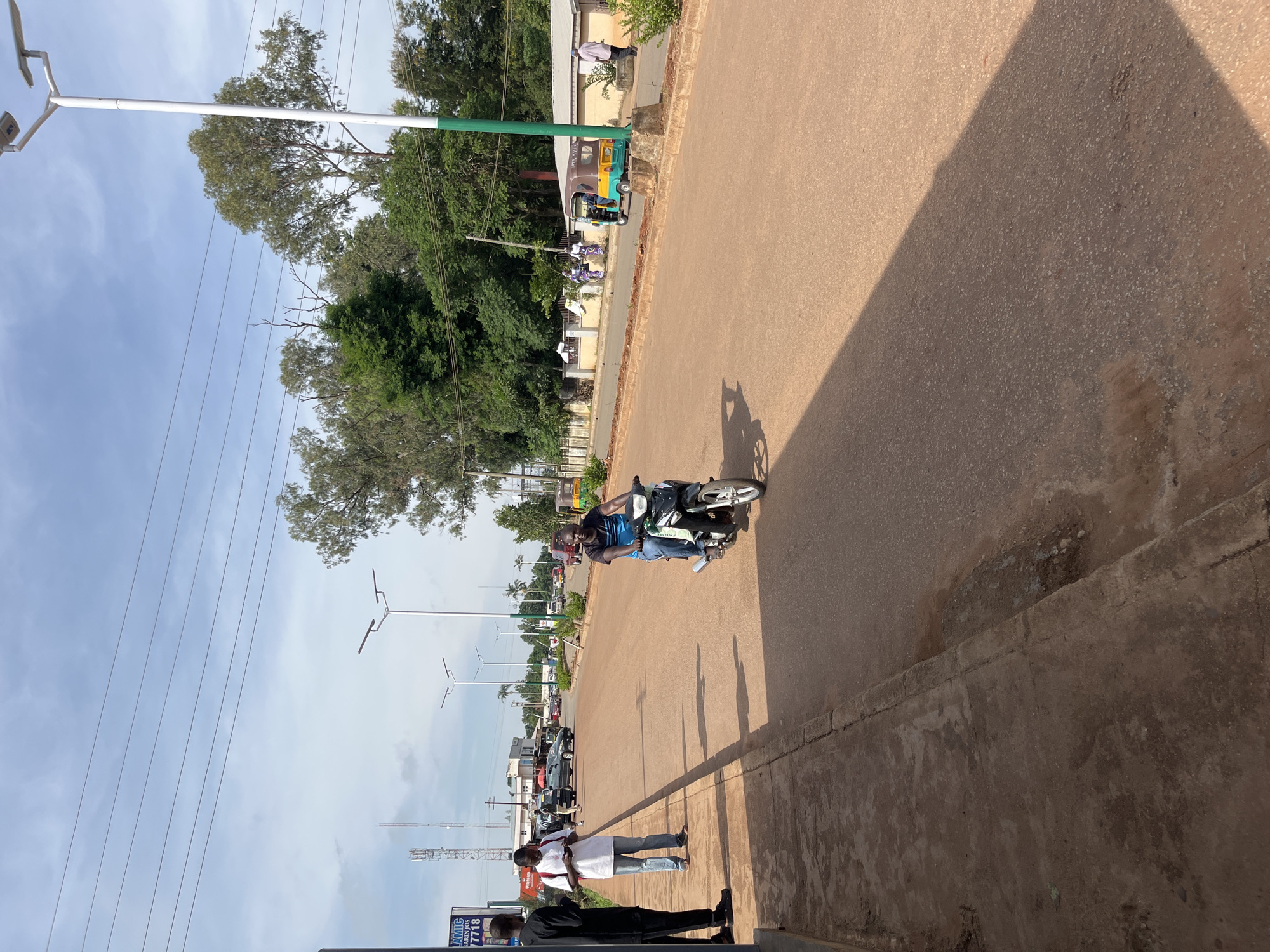 A sunny day in Bukuru, Nigeria, the sky is partly cloudy with a few scattered clouds. The scene is a wide, paved road with a reddish-brown surface, likely made of laterite or asphalt mixed with laterite. On the left side of the road, there is a concrete sidewalk with a few people standing and walking. A tall man in a black outfit stands on the sidewalk, his hands clasped in front of him. Next to him, a man in a white shirt and blue jeans is looking at his phone, and another man in a blue and black striped shirt is riding a black motorcycle down the road. The motorcycle has a green trim on the front and appears to be moving at a moderate speed. Further down the road, a few cars are visible, including a white SUV and a dark-colored sedan. A colorful tricycle, yellow, green, and brown, is parked on the right side of the road near a building. Trees line the background on both sides of the road, with a few tall, slender streetlights interspersed. The overall impression is one of a typical day in a bustling Nigerian city, with traffic, pedestrians, and everyday life unfolding.