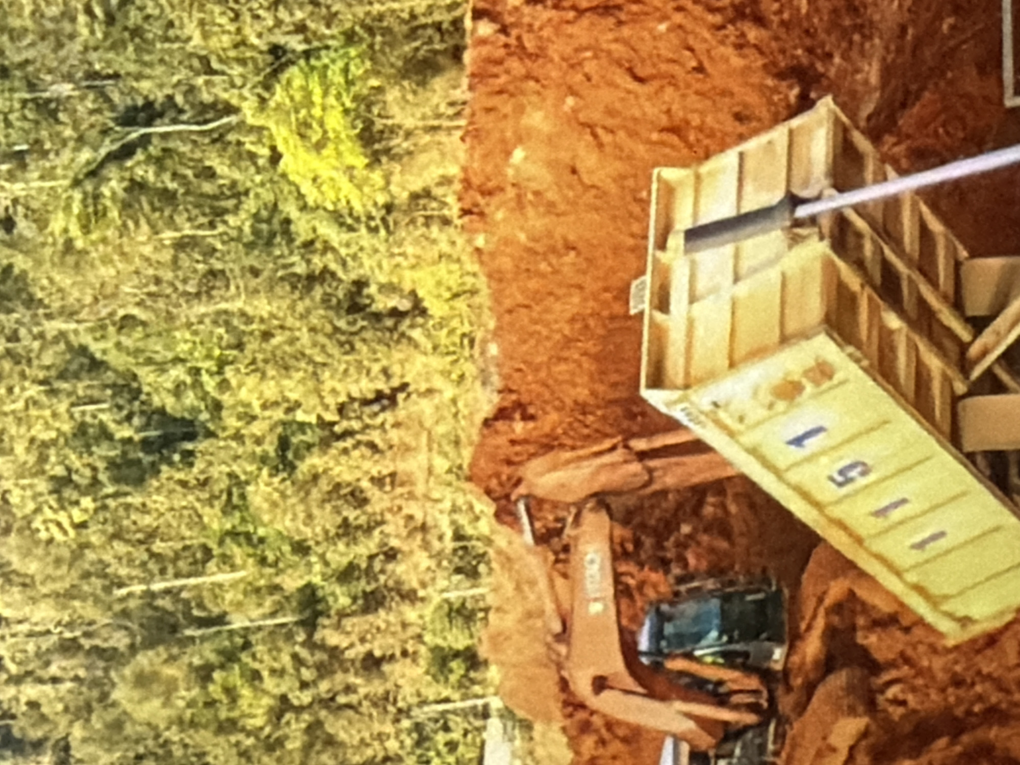 An excavator is visible with its arm extended towards a pile of dirt. The excavator has the brand name "HITACHI" visible on its arm. To the right of the excavator, a yellow dump truck bed is tilted upwards, displaying the numbers "1151" on its side. The background consists of a densely wooded hillside with trees in various shades of green and yellow. The ground appears to be a construction site with red dirt.