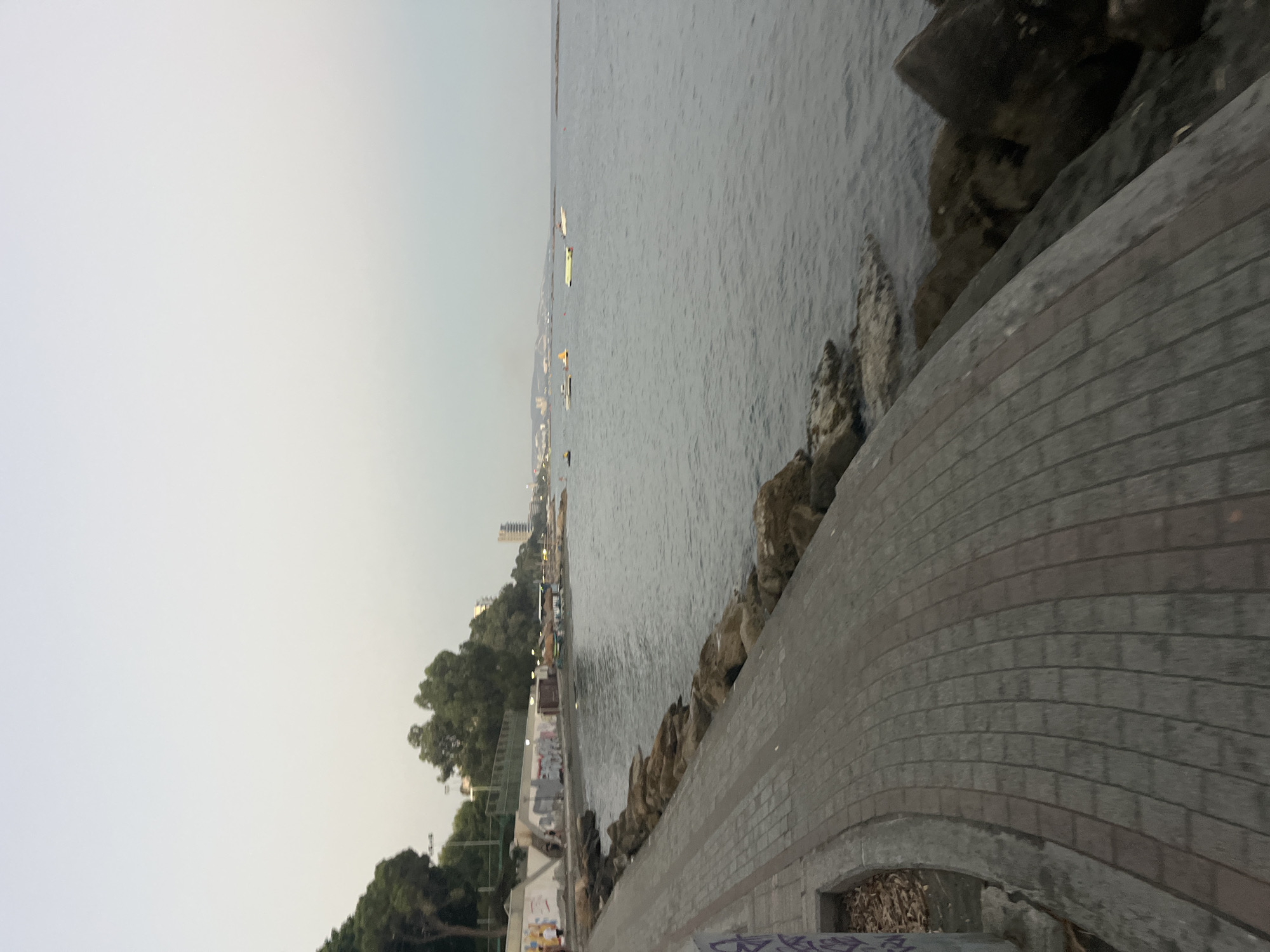 The image depicts a paved walkway alongside the sea in Agios Tychona Community, Cyprus. The walkway is constructed with rectangular paving stones and curves to the left, bordered by a low stone wall on the right. Beyond the wall, large rocks and boulders are visible along the shoreline. The sea is calm, with several small boats and what appear to be inflatable watercraft scattered on its surface. In the background, there is a treeline with scattered buildings and some taller structures, suggesting a coastal urban or semi-urban environment. The sky is overcast and the lighting suggests it is either early morning or late evening. The overall scene is one of a seaside promenade.
